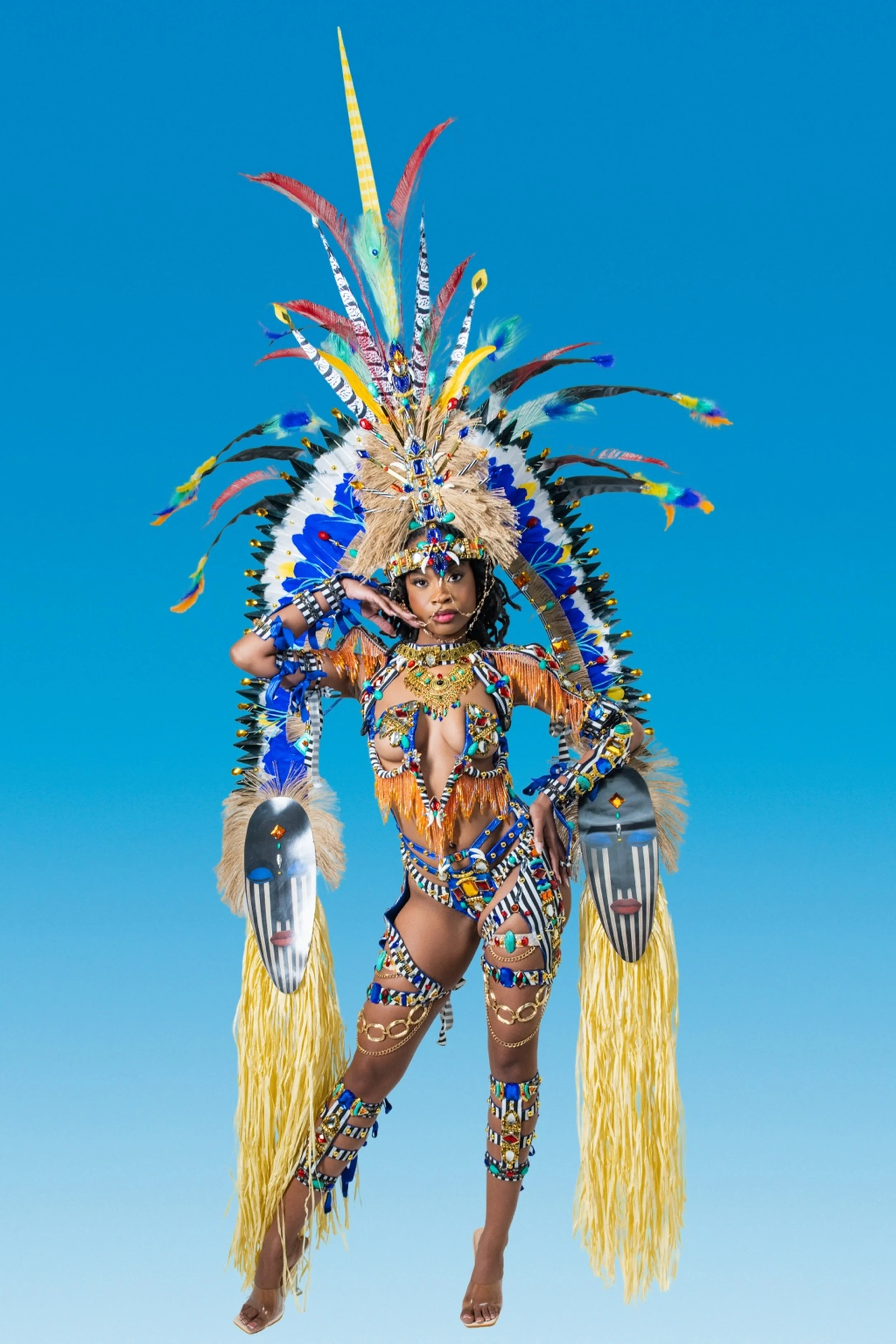 A woman dressed in a colorful, elaborate costume with feathers, beads, and masks against a blue background.