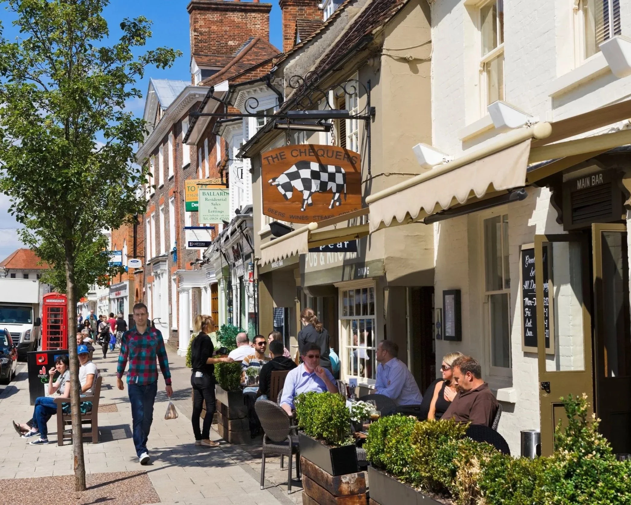 Marlow High Street near the E J Education school premises. People sitting outside a pub / restaurant on this lively street, with pedestrians walking and storefronts in the background under a bright blue sky.