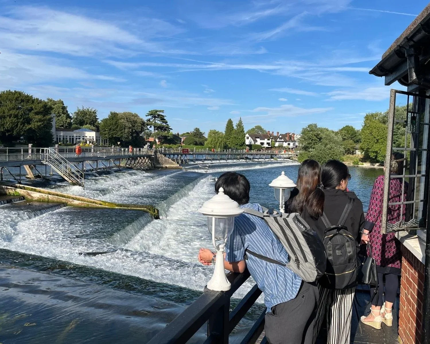 E J Education students enjoying the the view of the River Thames and Marlow Weir with water flowing over the edges from the balcony of the Compleat Angler Hotel on a sunny summer's day.