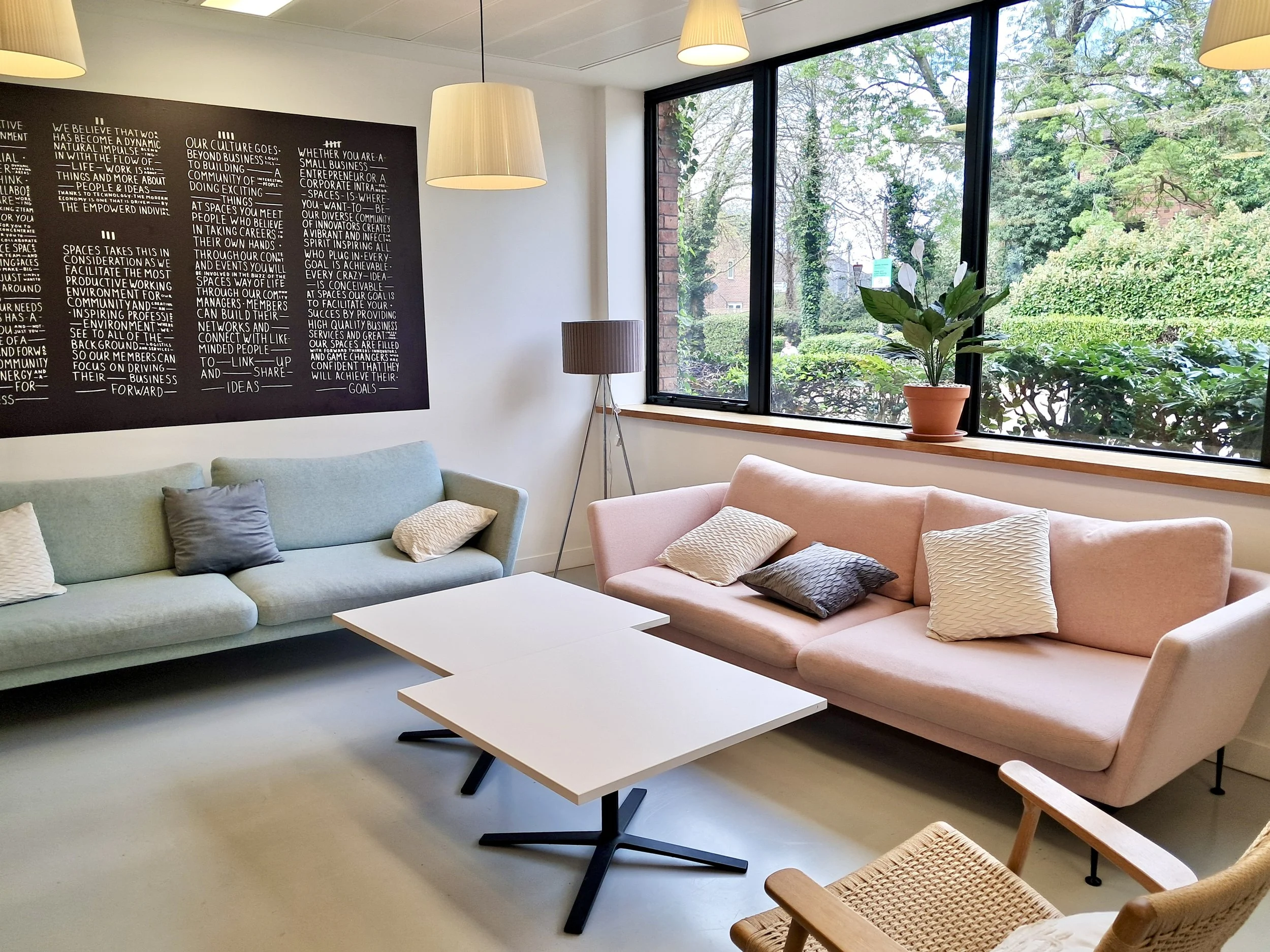 A cozy break-out area at E J Education's premises in Marlow with pastel-coloured sofas, white coffee tables, a floor lamp, and a large window showing trees outside. There's a black chalkboard with white text and a potted plant on the windowsill.