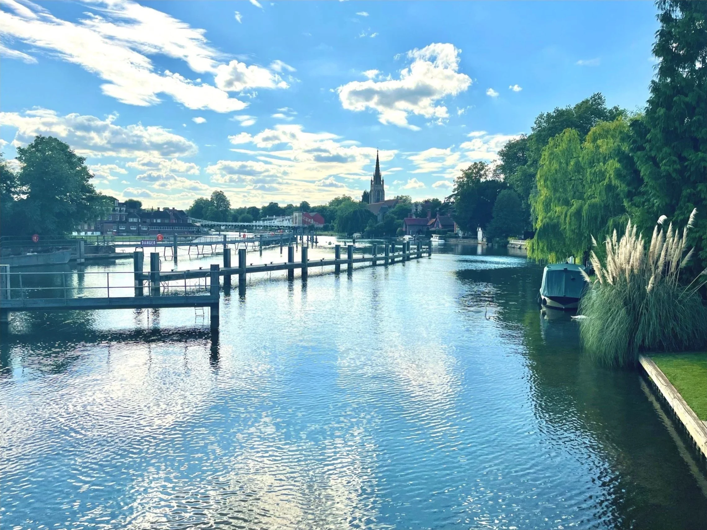 A scenic view of the River Thames in Marlow with trees on both sides, a boat docked on the right, and a church steeple in the distance under a partly cloudy sky.