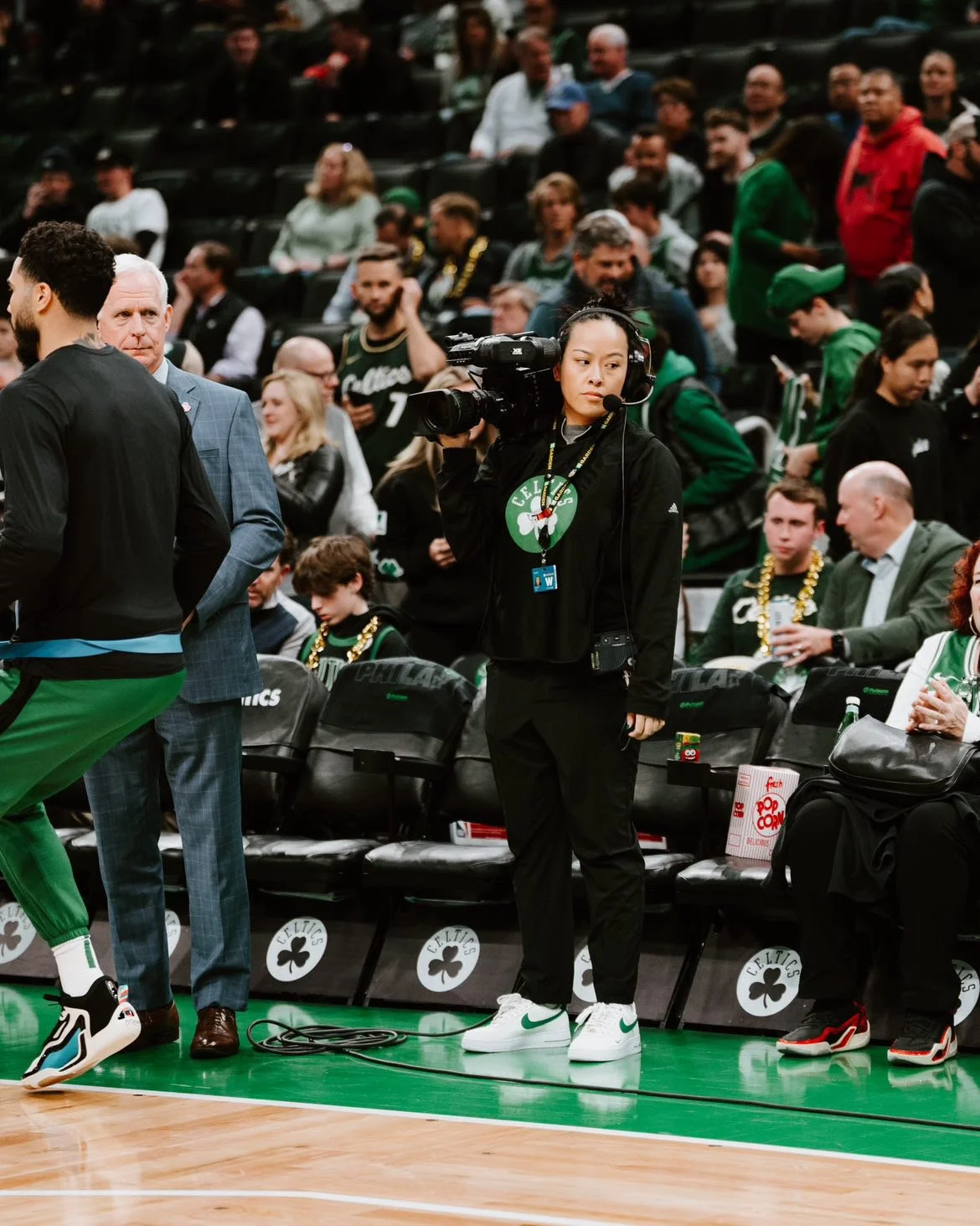 A woman wearing a black Celtics hoodie, black pants, and white sneakers with green Nike swoosh is holding a large video camera and standing near empty seats at a basketball game. The background shows many spectators, some wearing Celtics apparel and jewelry, in the stands of the arena.