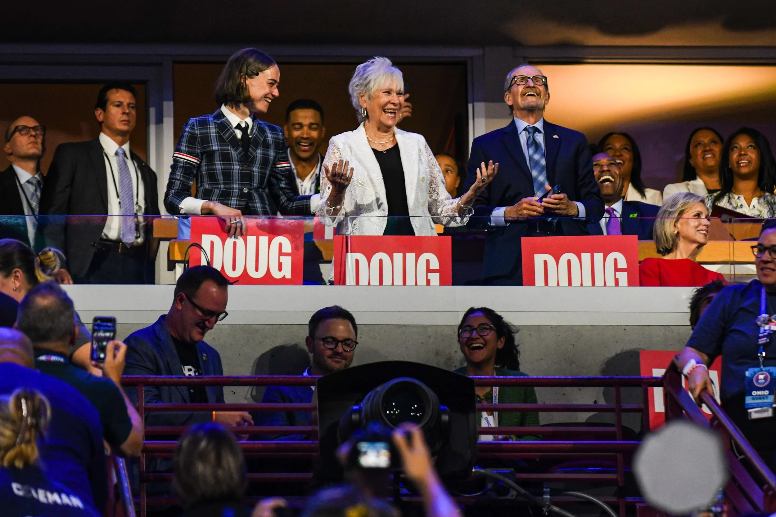 A group of people on a stage at a political event, with three individuals at the front behind signs labeled 'Doug'. The woman in the center has gray hair, is smiling, and has her hands raised. The man to her right is wearing glasses and a suit, also