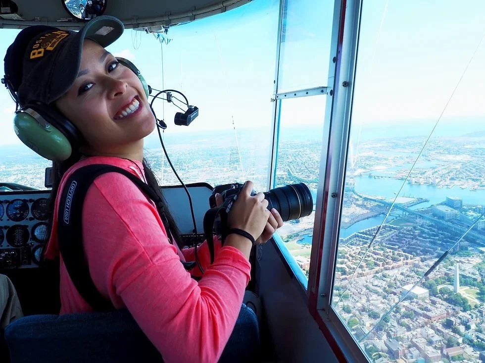 A woman smiling and holding a camera inside an aircraft cockpit, looking out the window at a cityscape with a river and numerous buildings.