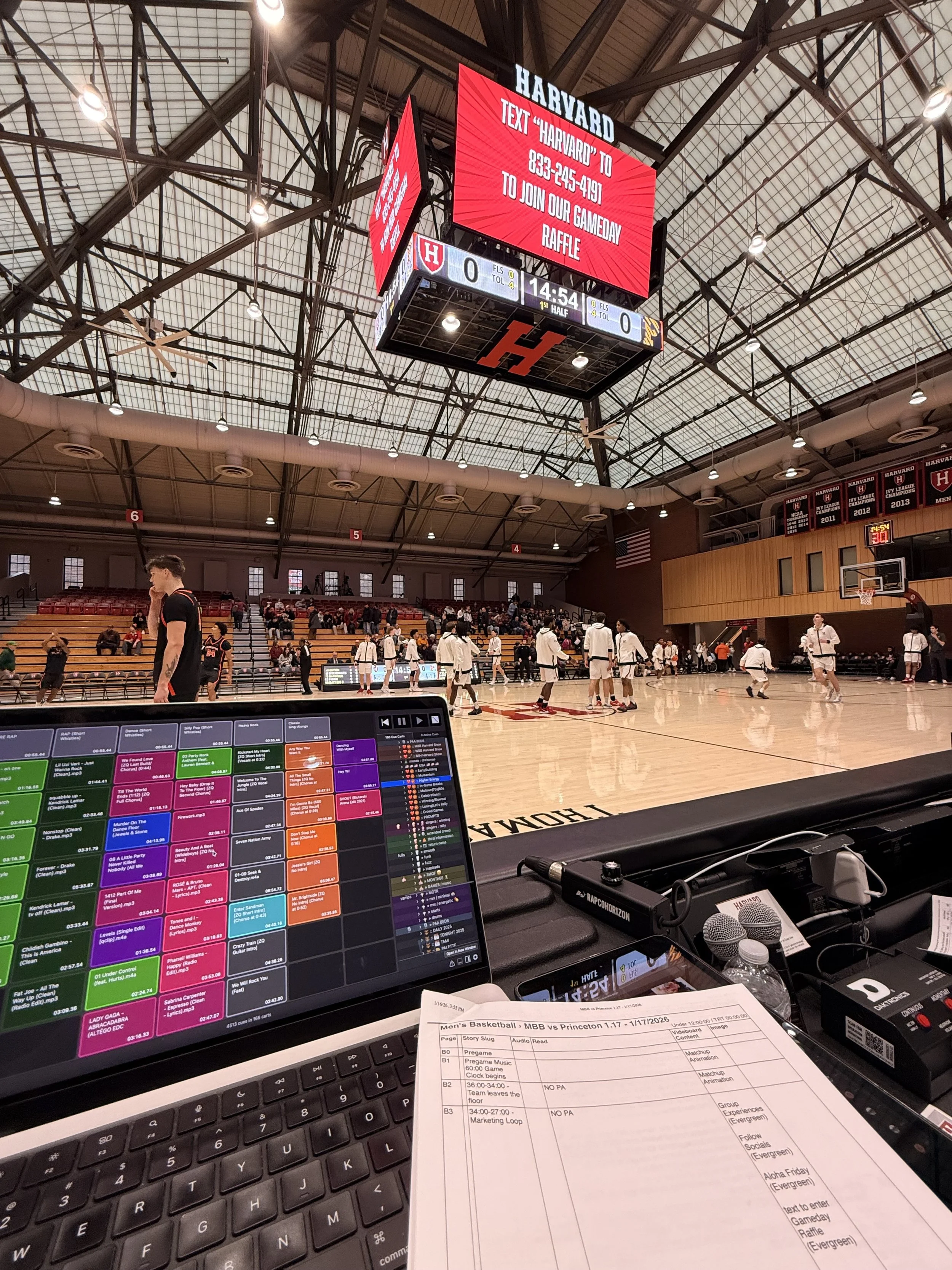Inside a basketball arena, people are warming up or practicing on the court. The view is fromannouncer's booth with a computer screen displaying a music playlist, a printed event schedule for men's basketball game, and various audio equipment.