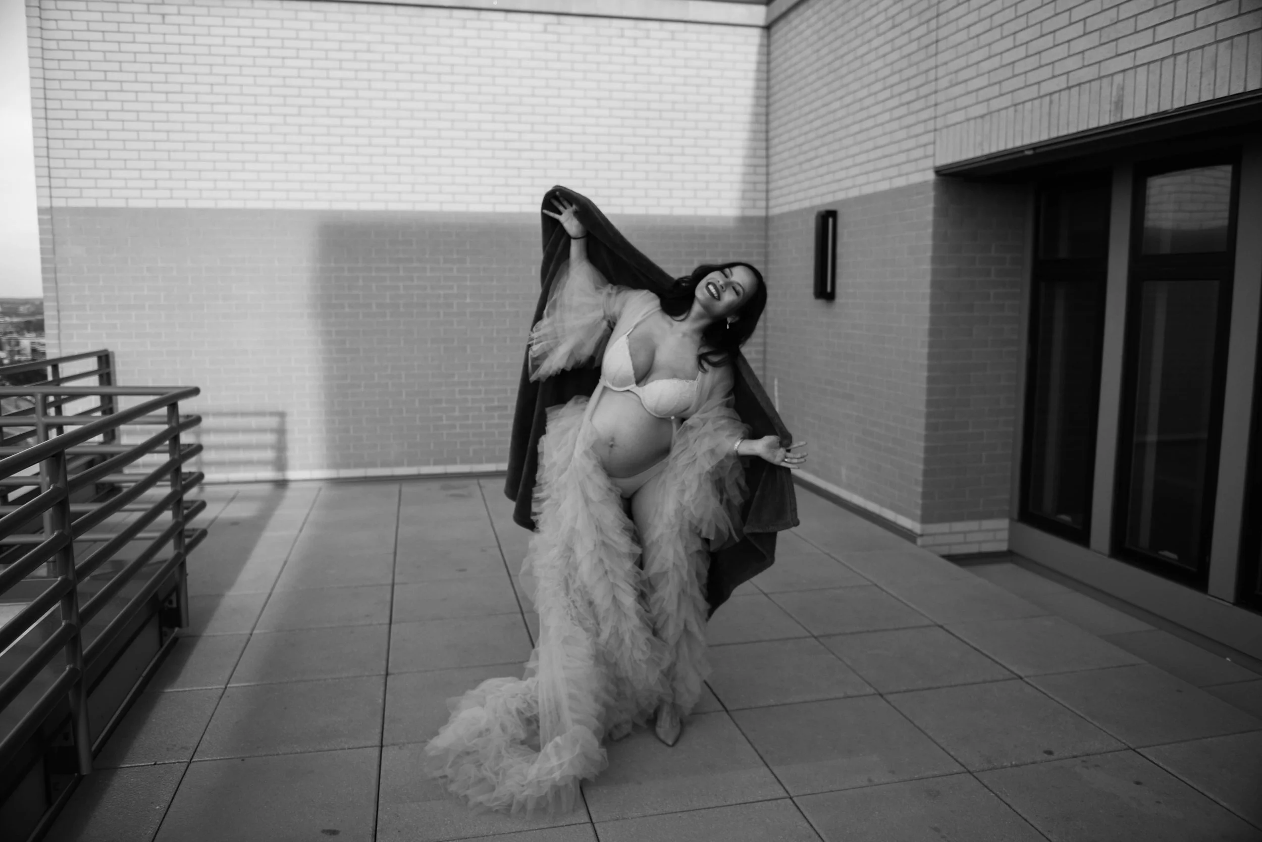 A pregnant woman in lingerie and a tulle skirt, posing on a balcony with brick walls and large windows, smiling and holding her hair, in black and white.