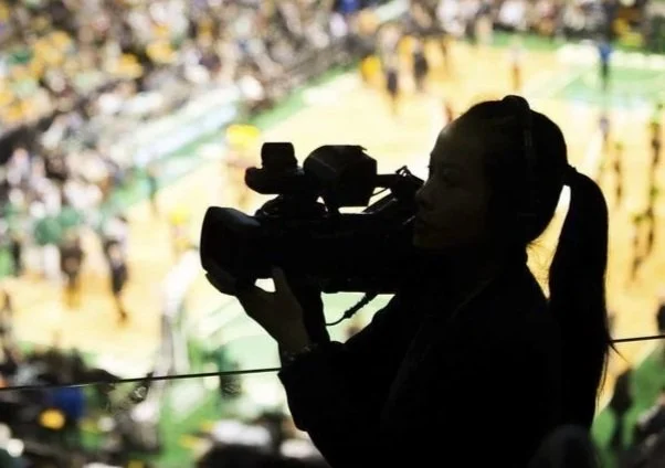 Silhouette of a female videographer filming at an event or sports game with a green field in the background.