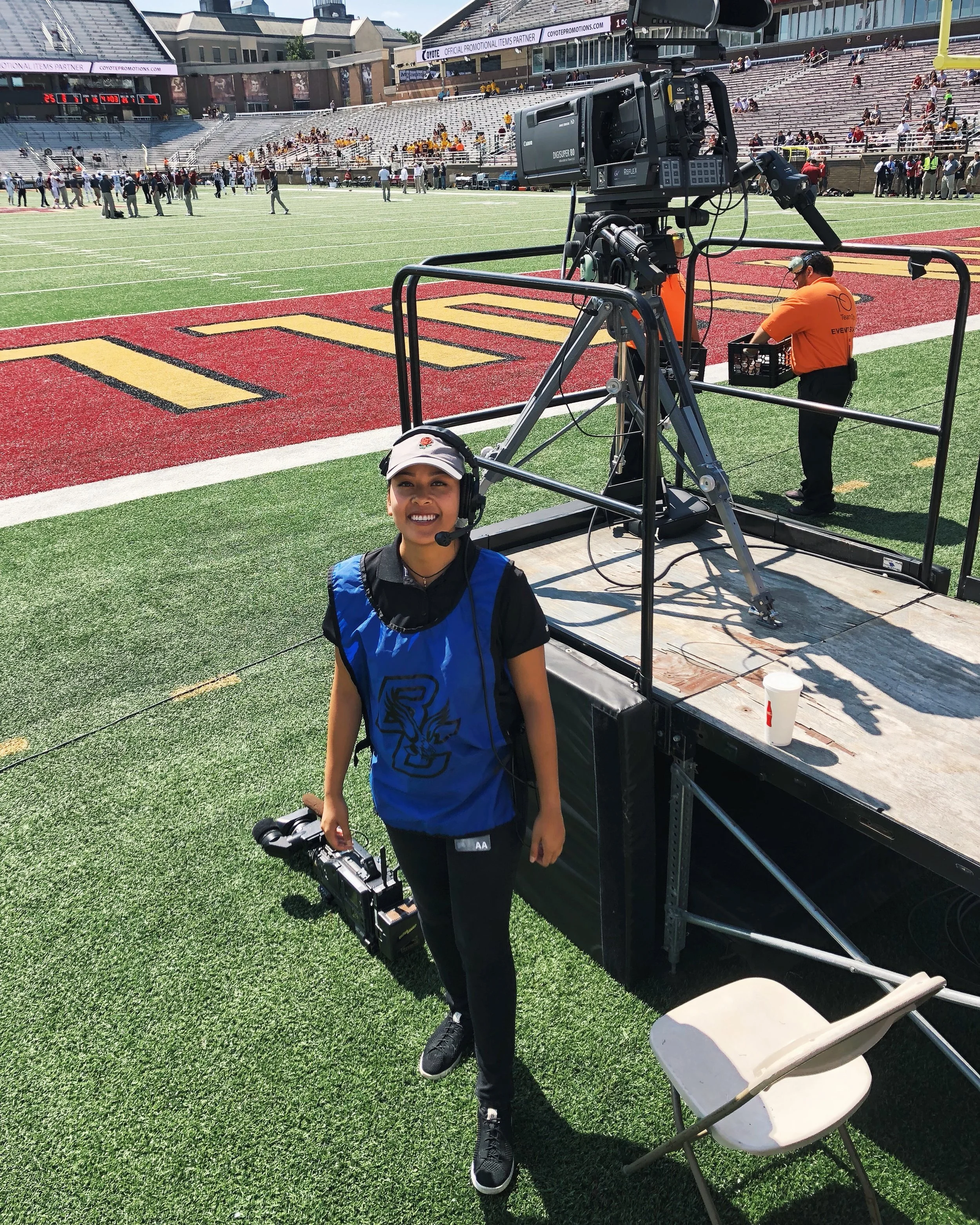 A woman standing on a football field near camera equipment and a person in an orange shirt. The woman is smiling and wearing headphones with a microphone, a blue vest, black pants, and a cap. She holds a camera in her right hand. The scene is taking place at a stadium with people in the background.