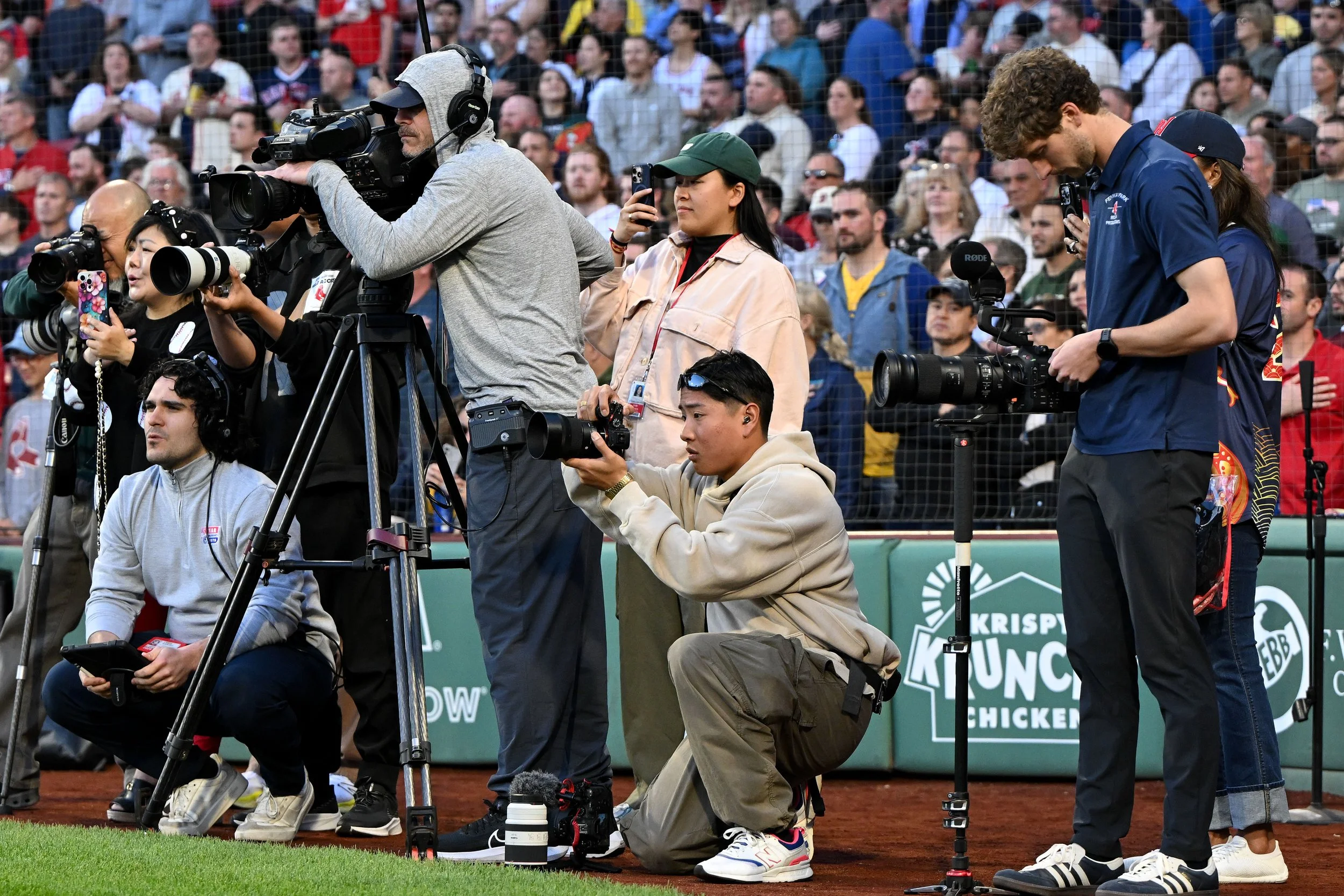 Multiple photographers and videographers are capturing an event on a field with a large crowd watching in the background.