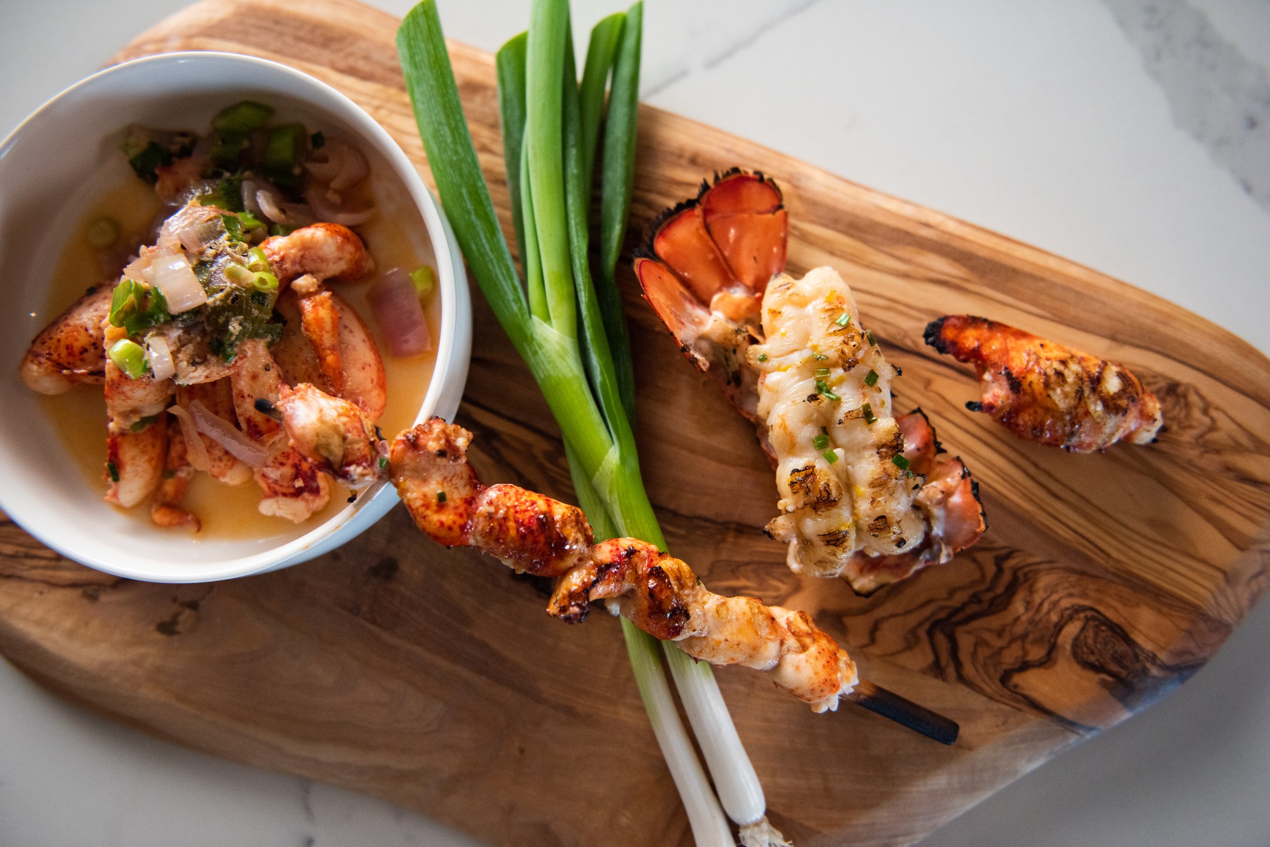 Grilled lobster tail, skewered grilled chicken, green onions, and a bowl of lobster meat salad on a wooden serving board.