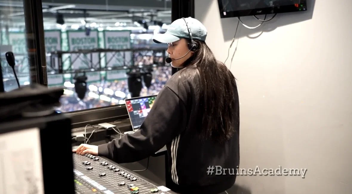 Female audio technician wearing a headset, operating broadcasting equipment in a control room overlooking a sports stadium.