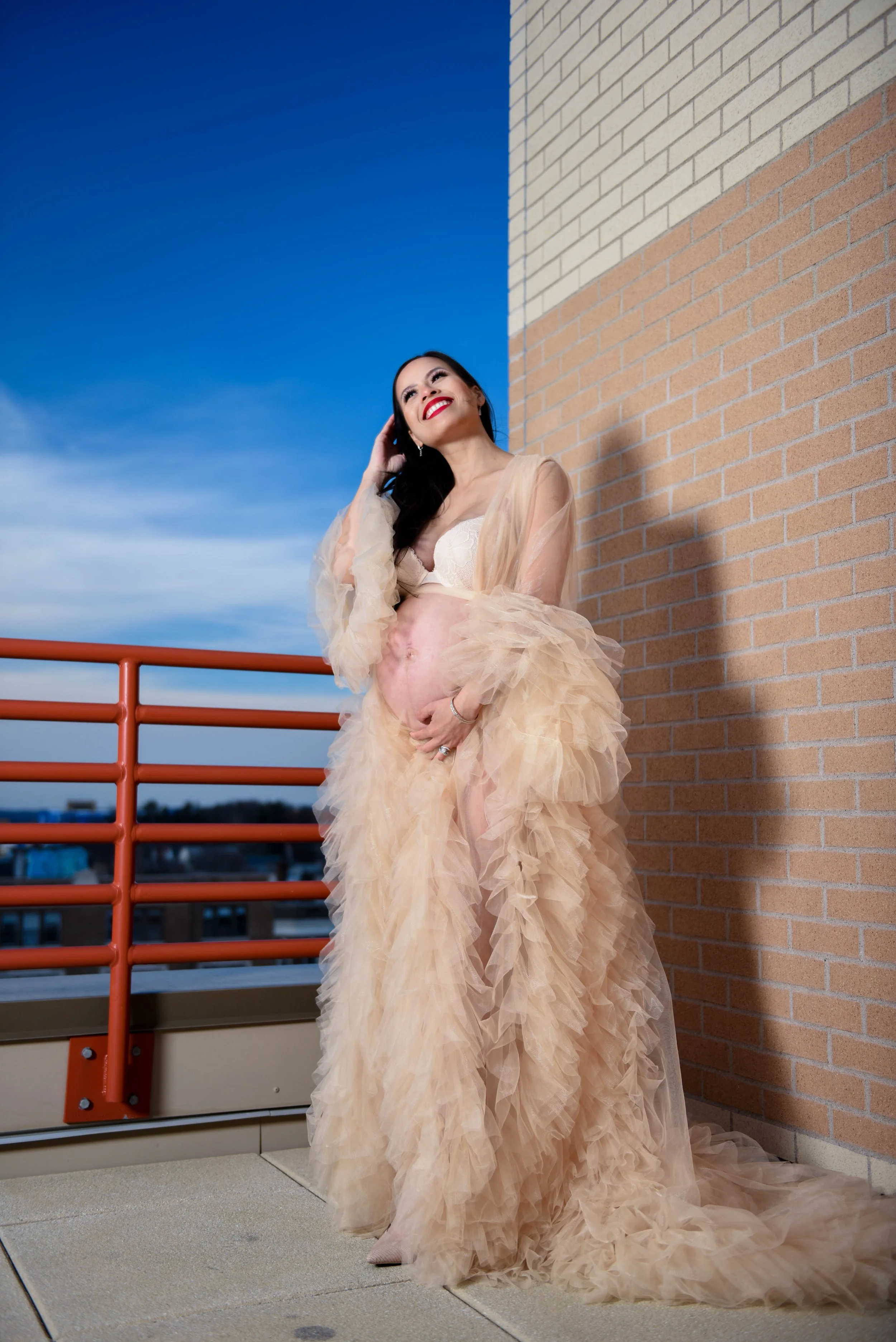 A smiling pregnant woman in a beige, ruffled gown standing outside against a brick wall, with a blue sky in the background.
