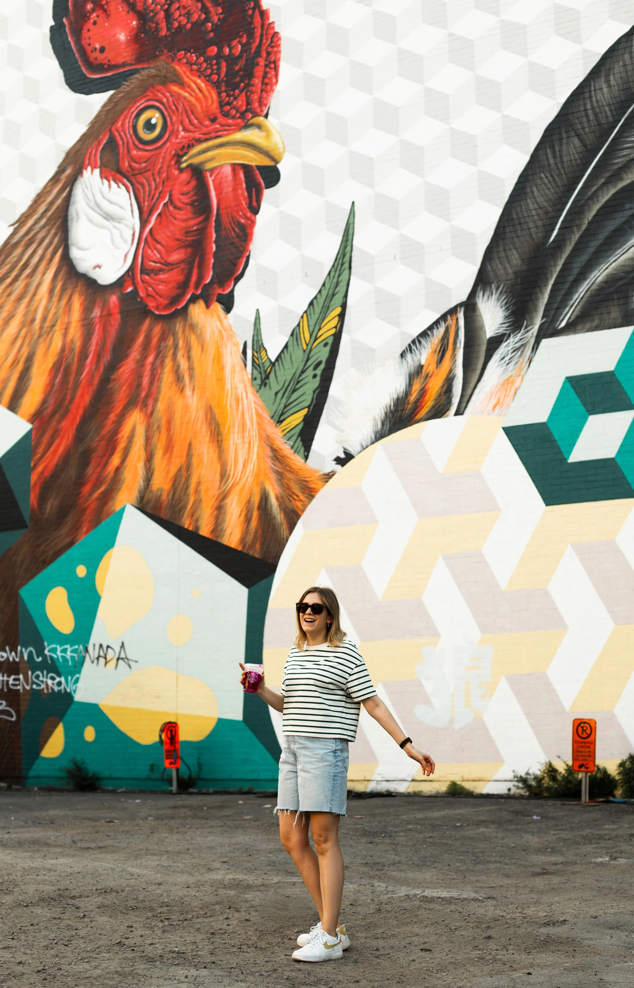 A woman with sunglasses and a striped shirt holding a drink standing in front of a large colorful mural of a rooster and other abstract elements.