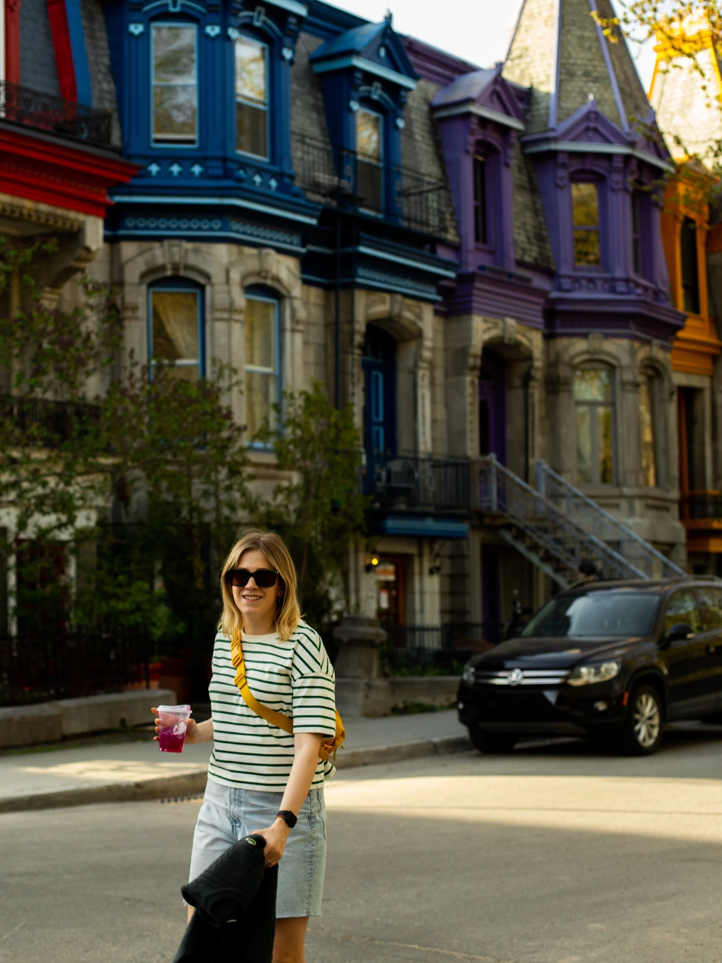 Woman with blond hair and sunglasses smiling on a city street, holding a drink and a hat, with colorful Victorian-style row houses in the background.