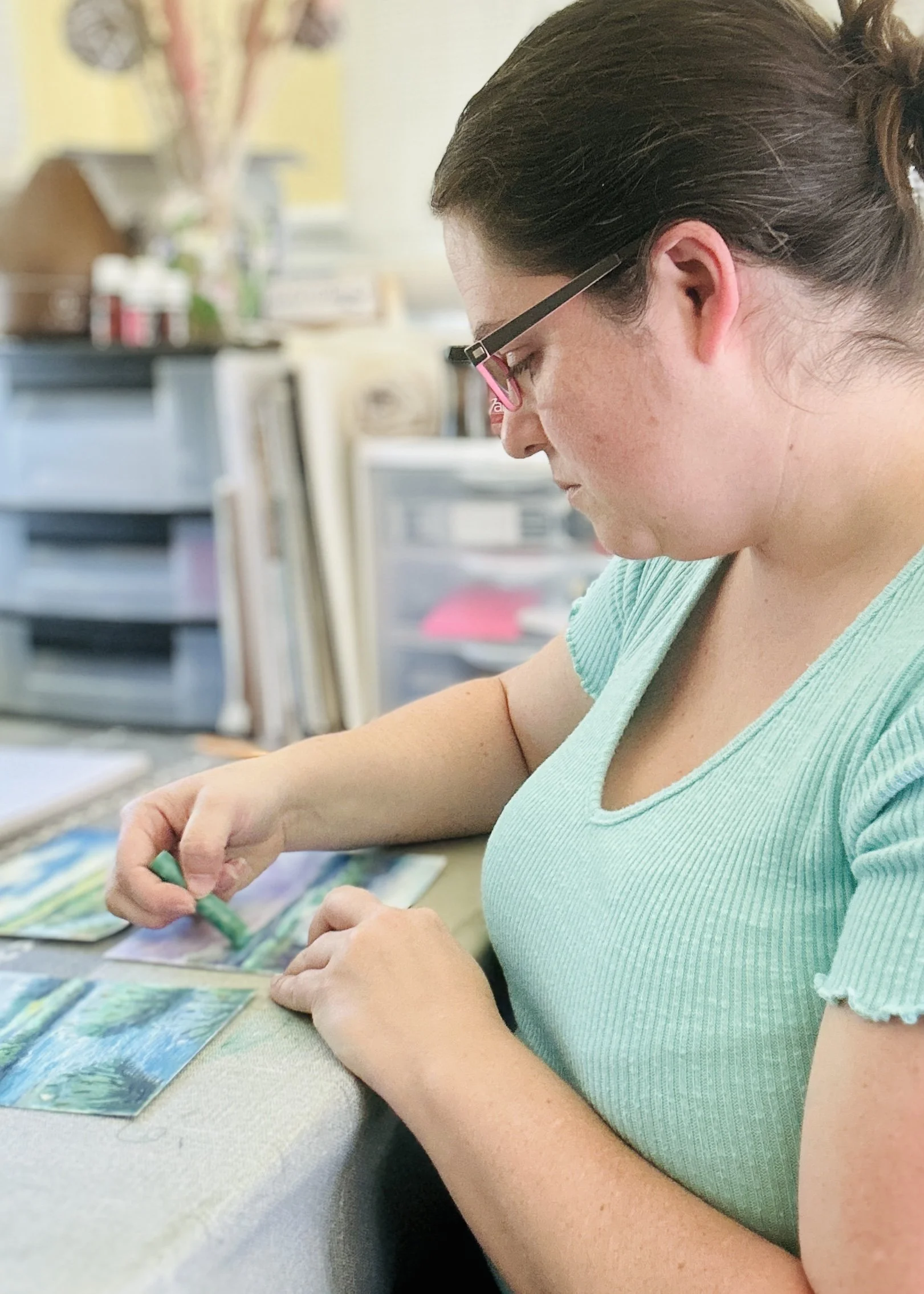 Artist Bek Harris in a turquoise shirt is sitting at a table in her studio, working on small art pieces.