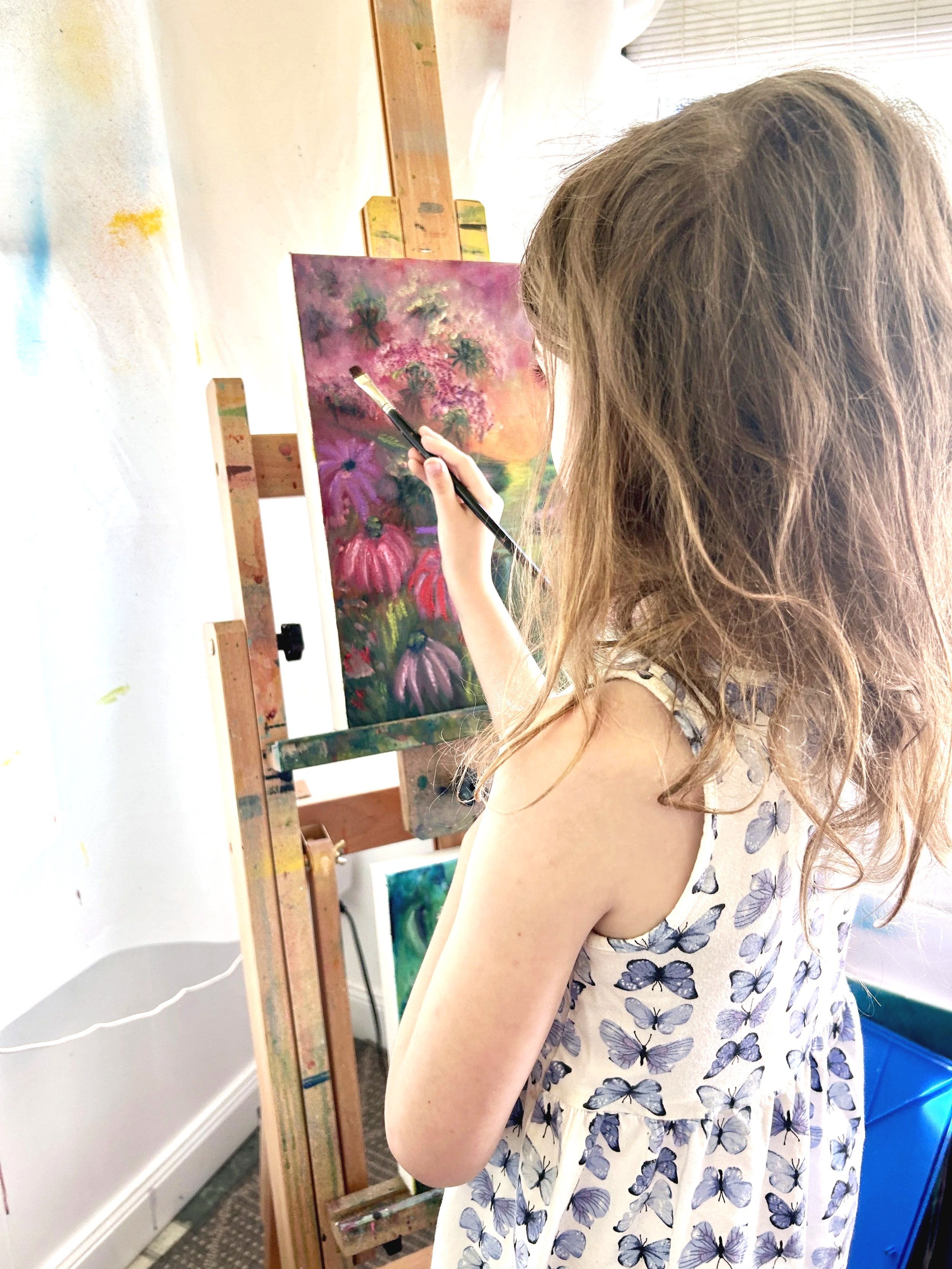 A person with light brown wavy hair, wearing a sleeveless dress with a butterfly pattern, is painting on a canvas with a brush in an art studio. There are paintings, a wooden easel, and art supplies in the background.