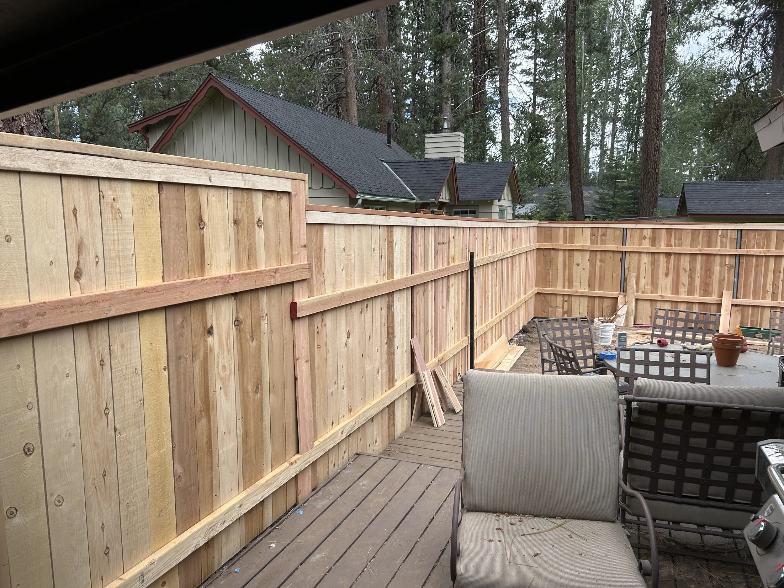 Backyard patio under construction with wooden fencing, patio furniture, and building materials, surrounded by trees and neighboring houses.