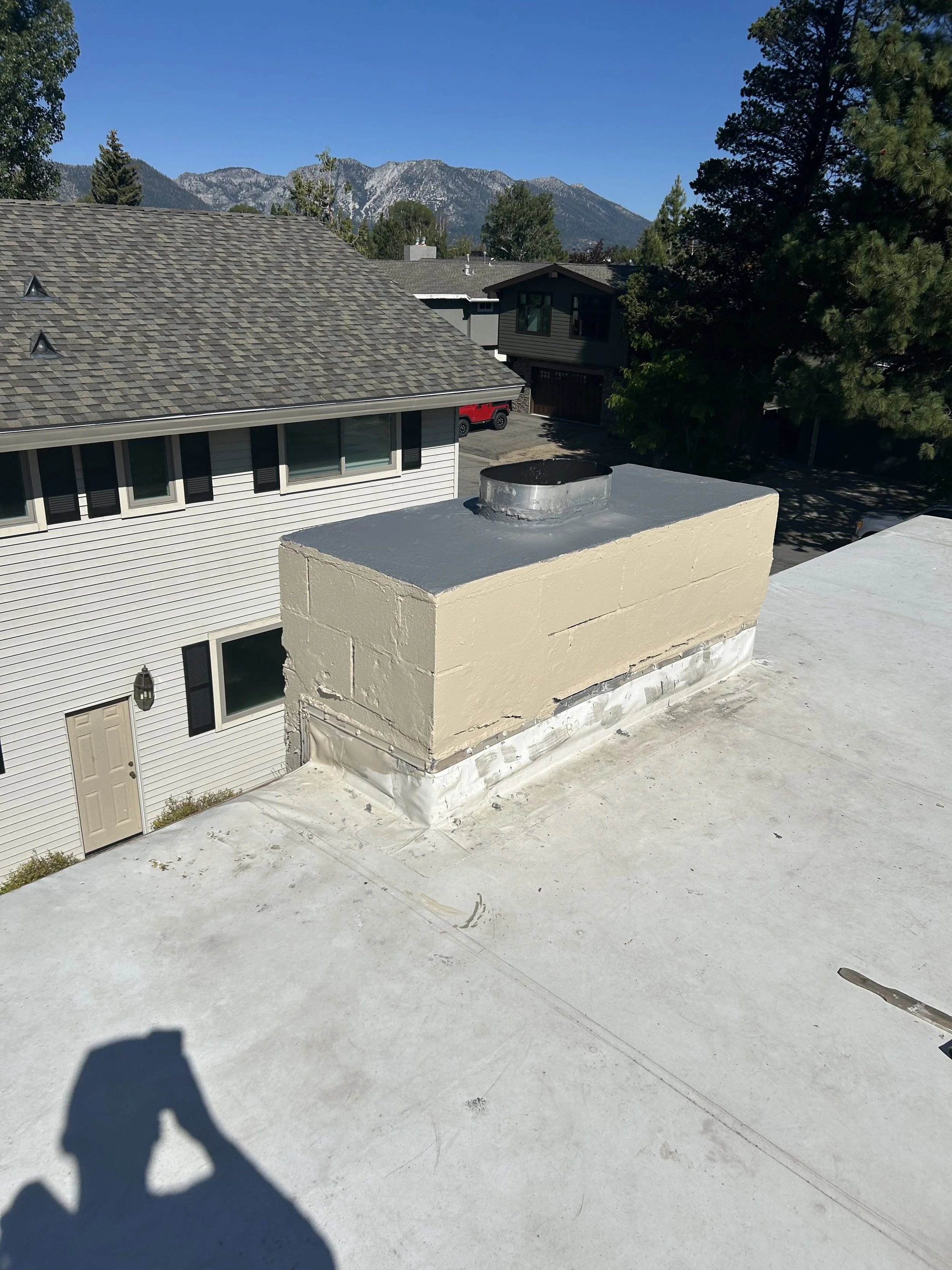 View of a rooftop chimney with a beige stucco exterior and a black metal cap, situated on a flat white roof. In the background, there are neighboring houses, trees, and distant mountains under a clear blue sky.
