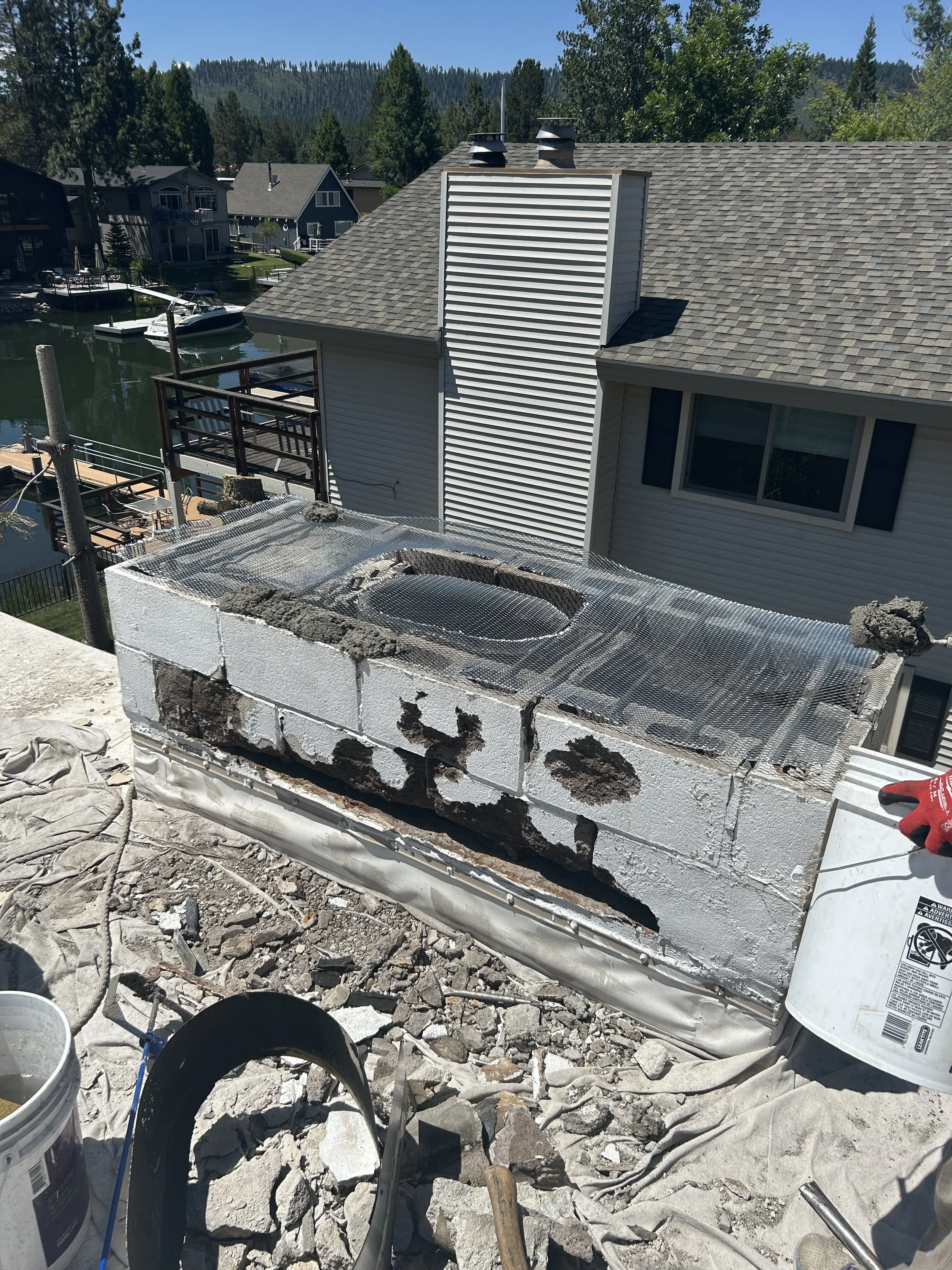 Construction worker repairing an air conditioning unit on a rooftop. The unit is partially damaged with visible concrete blocks and insulation. Residential houses, a body of water, boats, and trees are visible in the background.