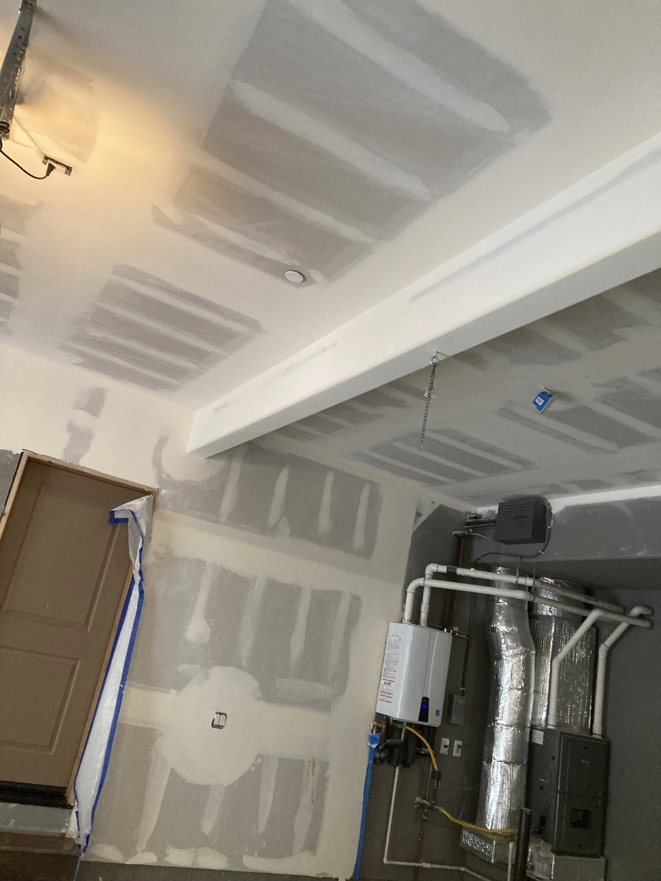 Interior view of a room under construction or renovation, with drywall walls and ceiling patch, a garage door, and visible HVAC ductwork and equipment.