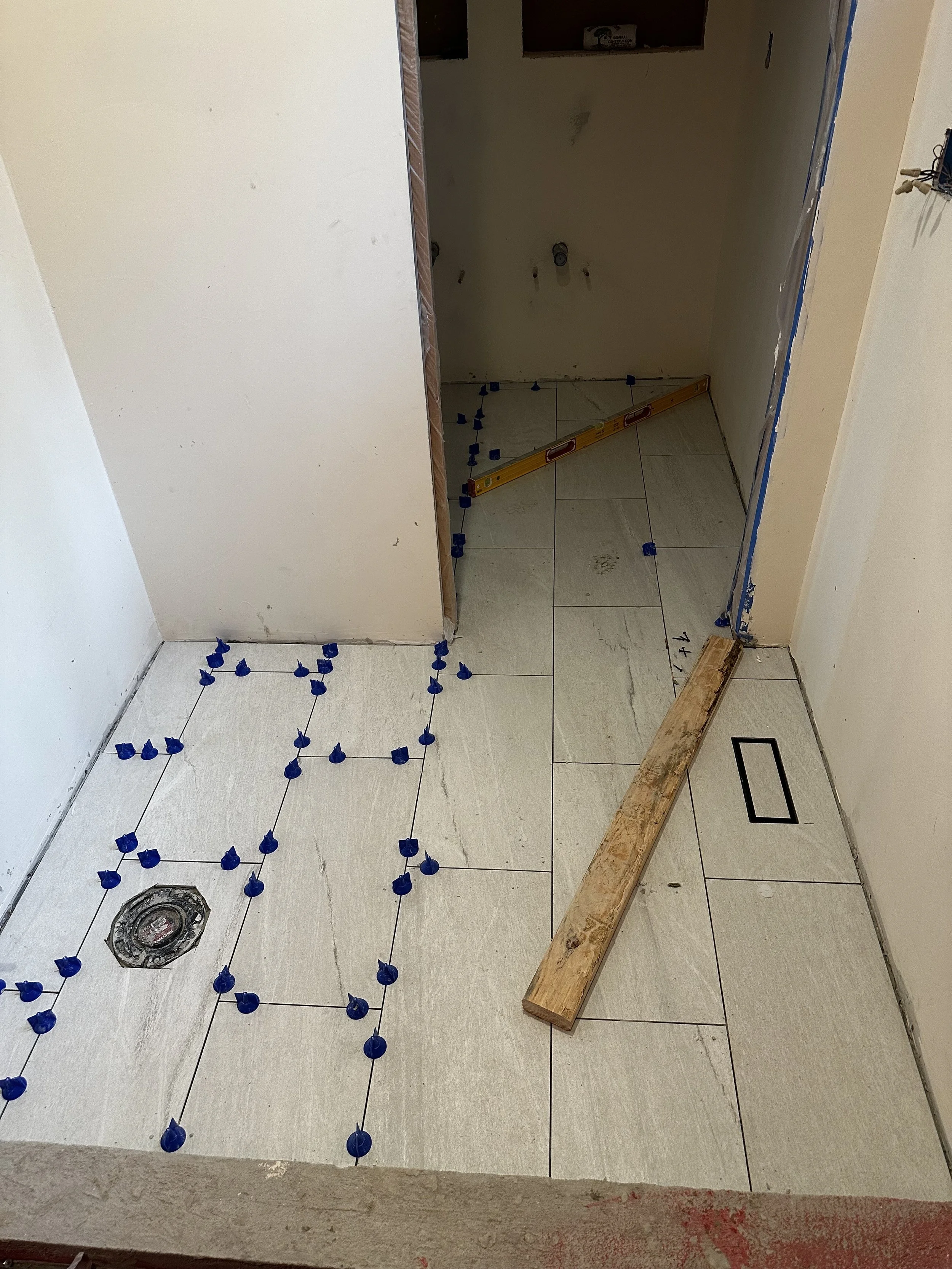 Bathroom under renovation with tiled floor, blue tile spacers, a level tool, and plumbing. Roots to Rooftops General Contractor
