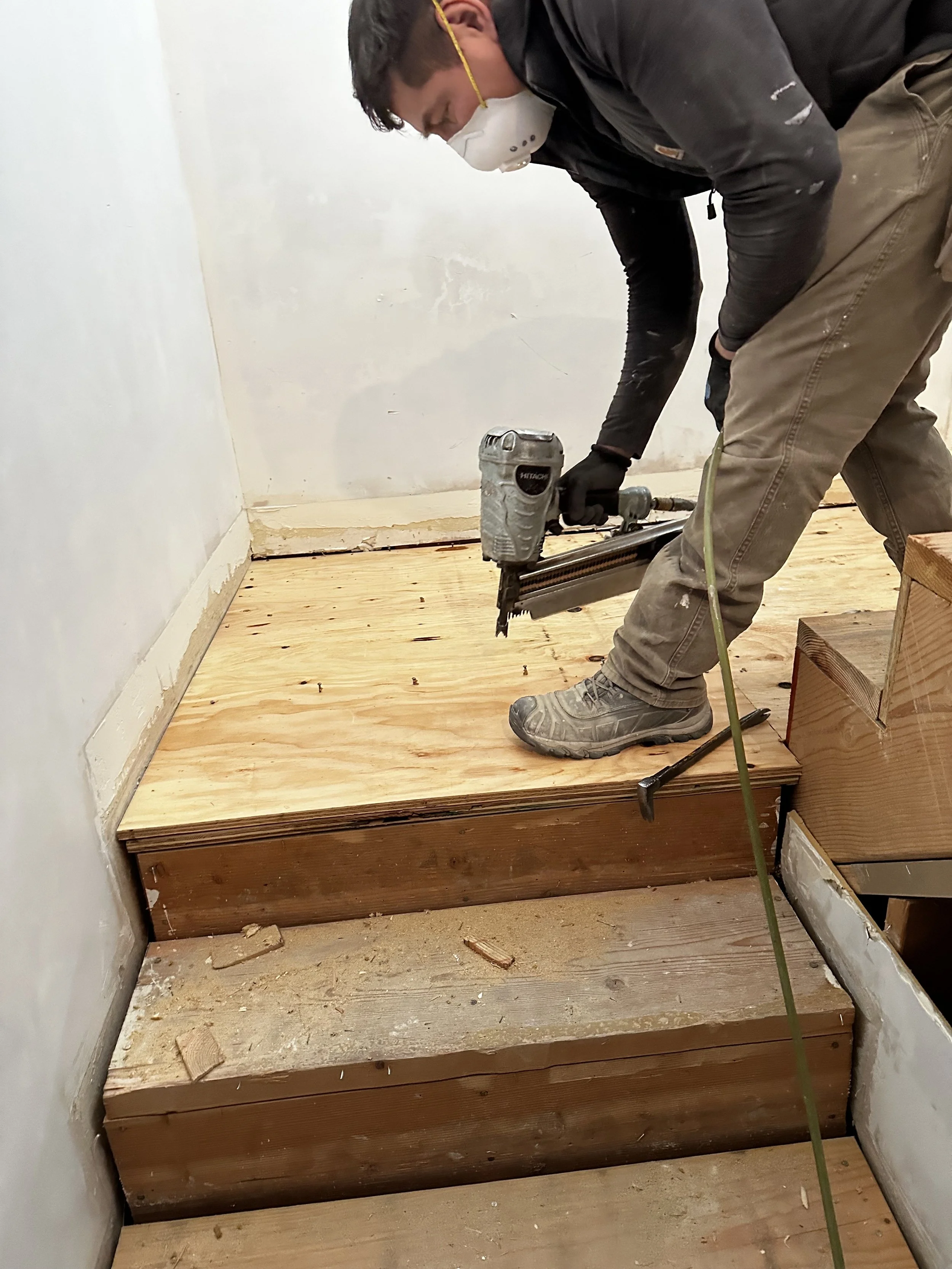 A person wearing a face mask, gloves, and work boots is using a nail gun to attach a plywood subfloor onto a staircase.
