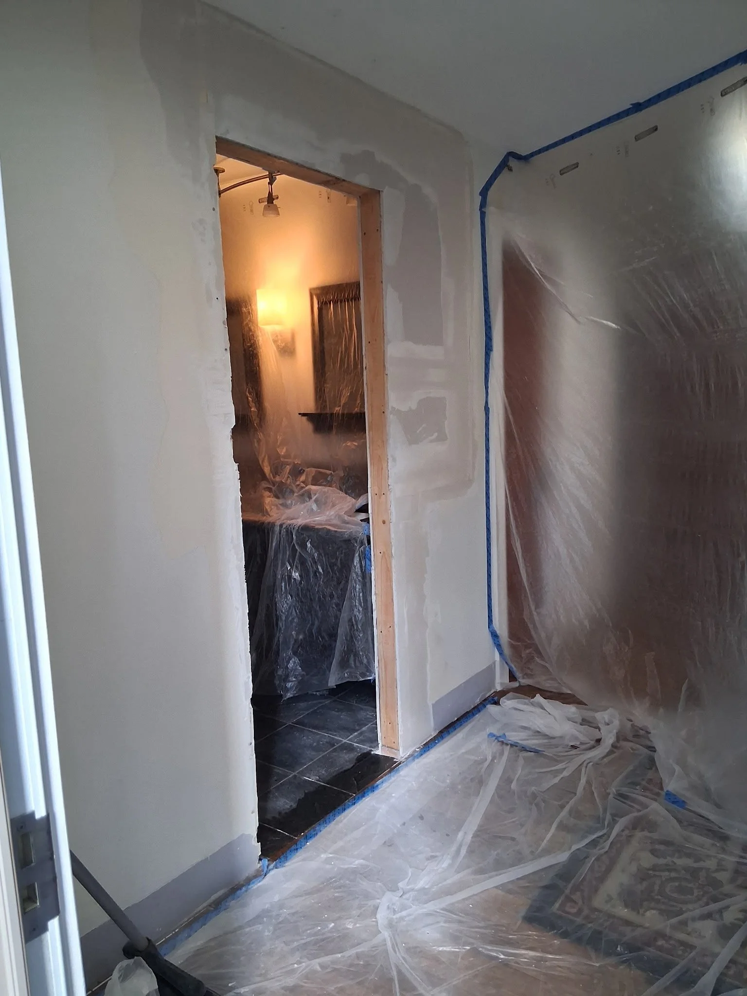 Interior room under renovation with plastic covering on the floor and walls, partially painted wall, doorway leading to another room with a lamp and wall art, construction supplies visible.
