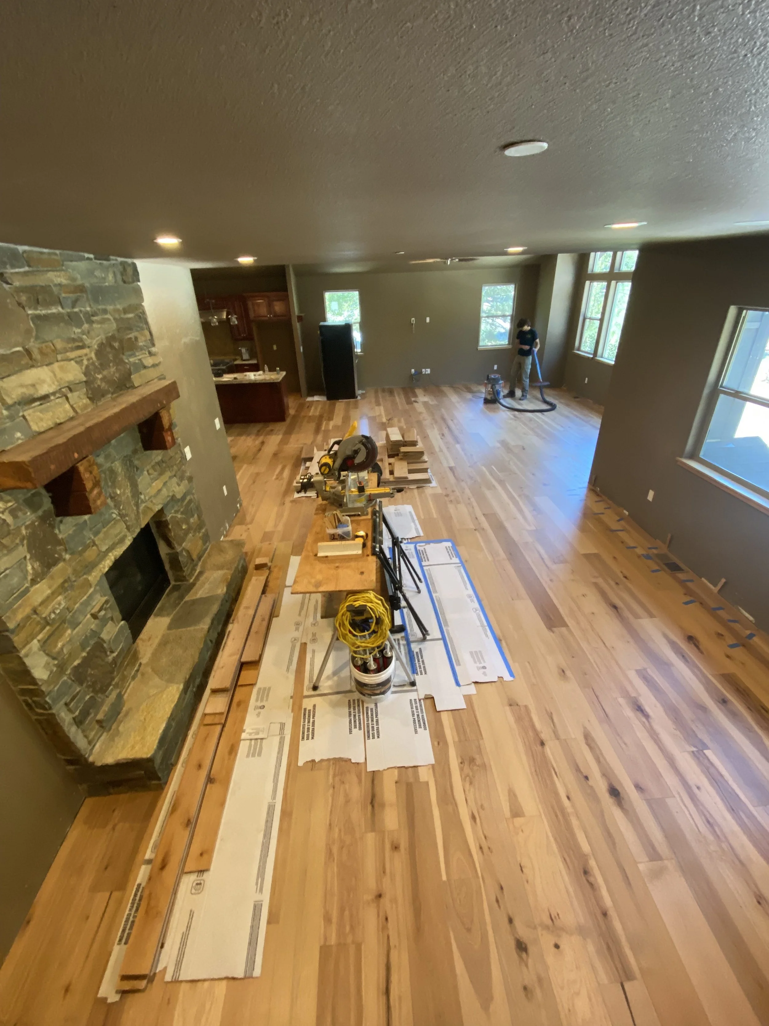 Living room under construction with tools and wood planks on the floor, a person working near the windows, and a stone fireplace on the left. Replace flooring hardwood, lvp, laminate.