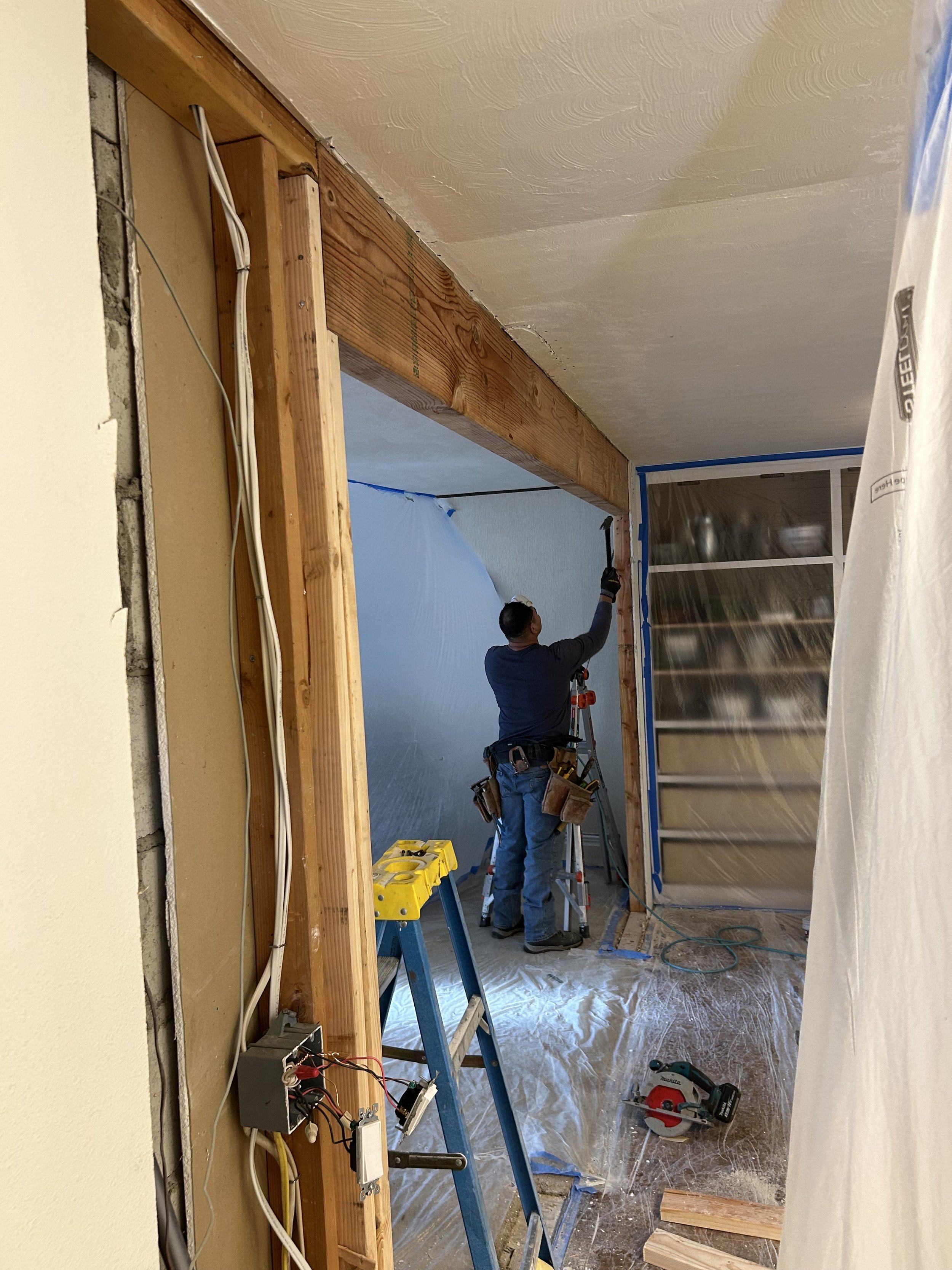 A worker is standing on a ladder, installing or repairing a wooden beam in a room under renovation. The room has plastic sheeting covering parts of it, and construction tools are visible on the floor and a nearby ladder.