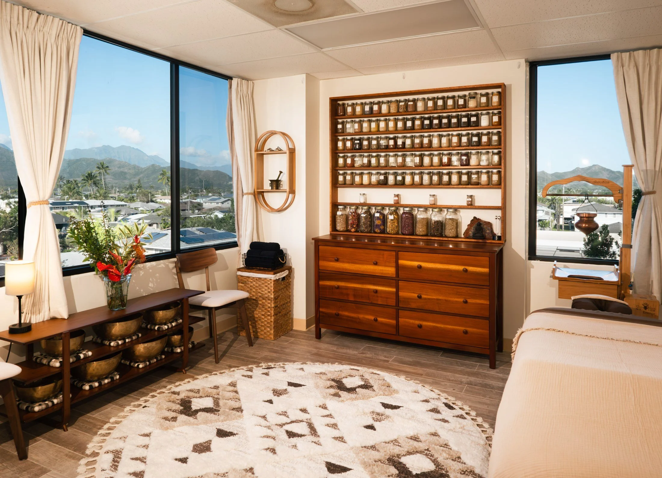 Ayurvedic treatment room in Kailua with herbal shelves, singing bowls, a massage table, and views of the Koʻolau mountains.