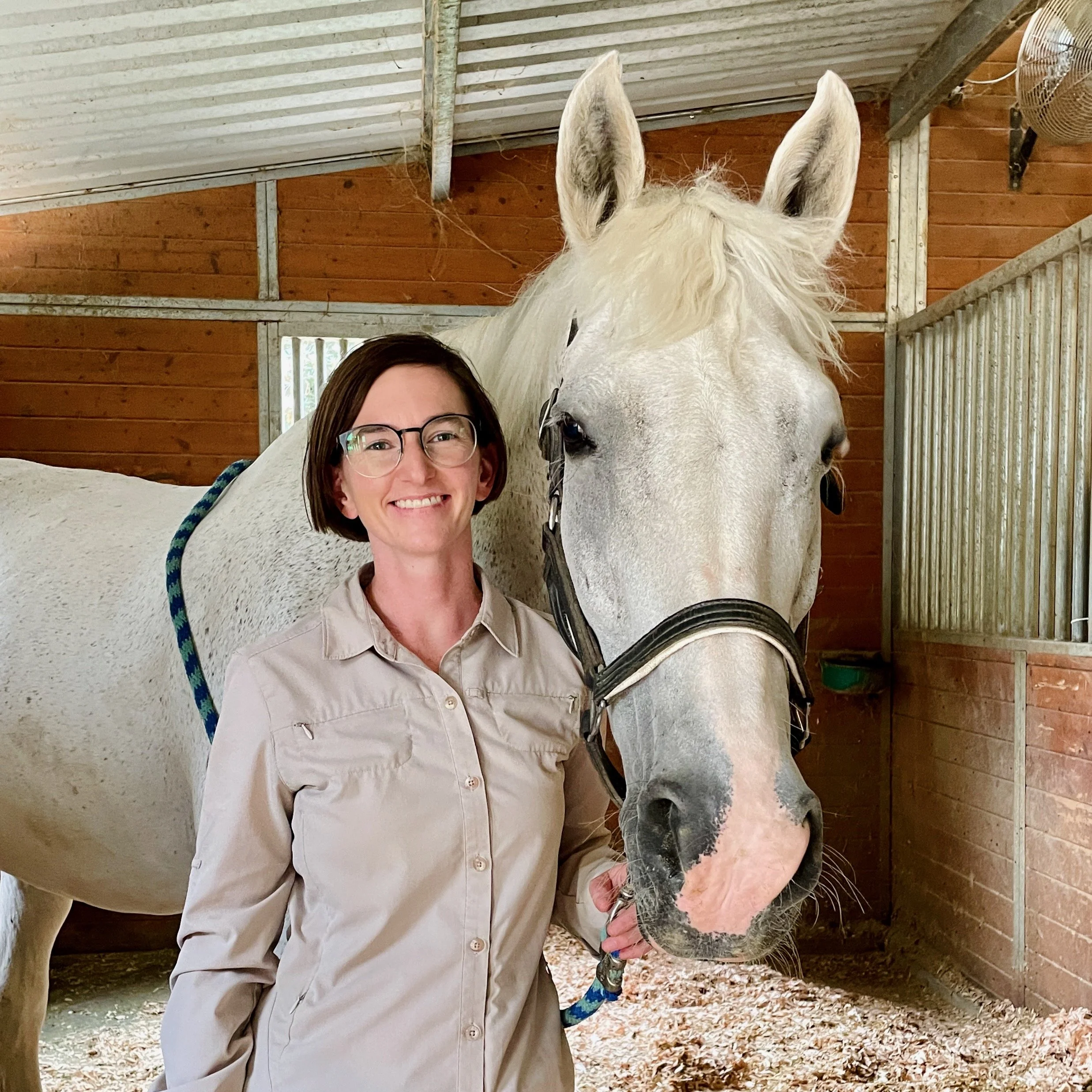 Smiling woman wearing glasses poses beside a white horse in a wooden stable.