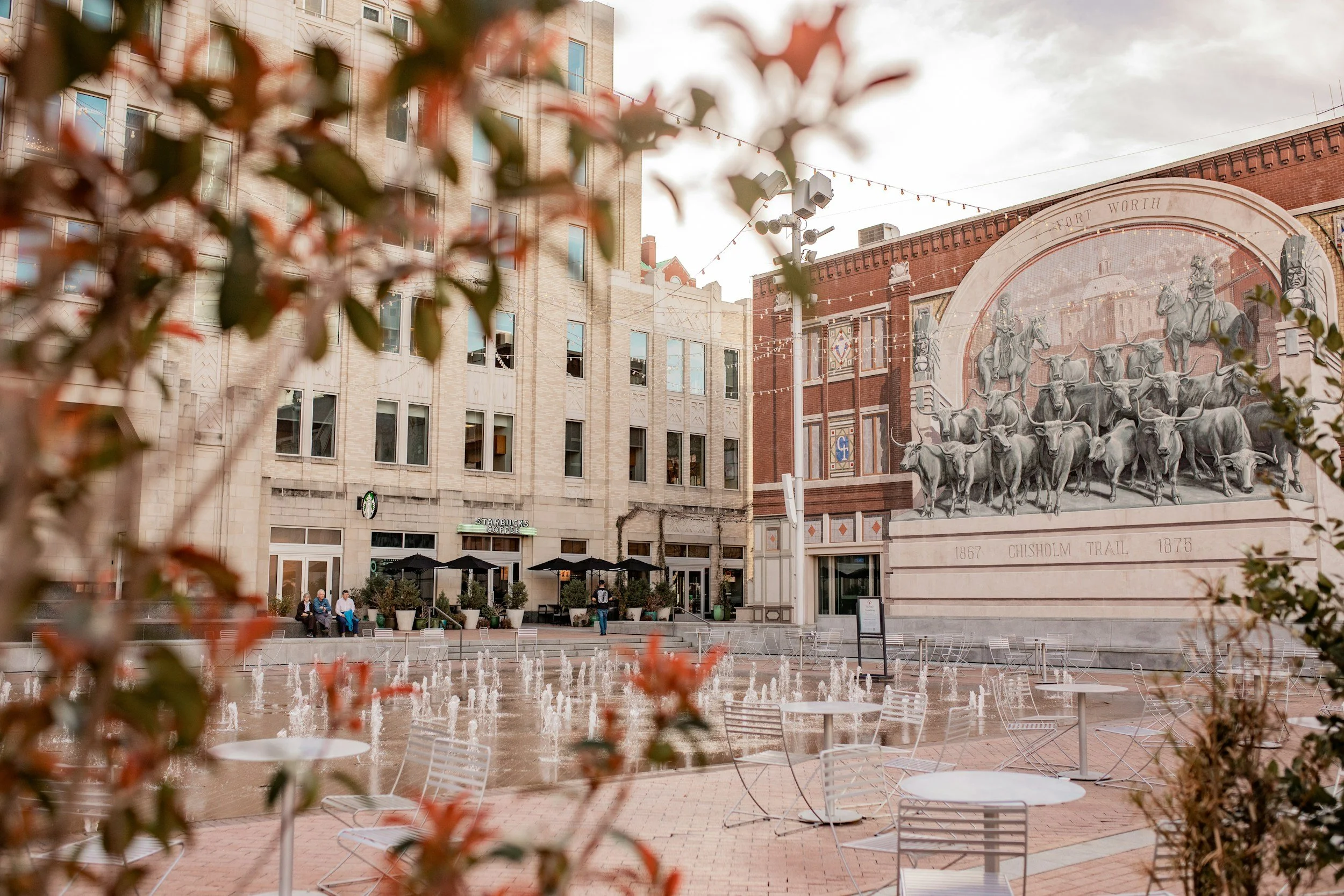 A city square with a fountain, outdoor tables, and chairs in front of a mural depicting herds of cattle, with a red brick building to the right and a beige stone building behind. Some people are sitting and walking, and there are string lights hanging over the area.