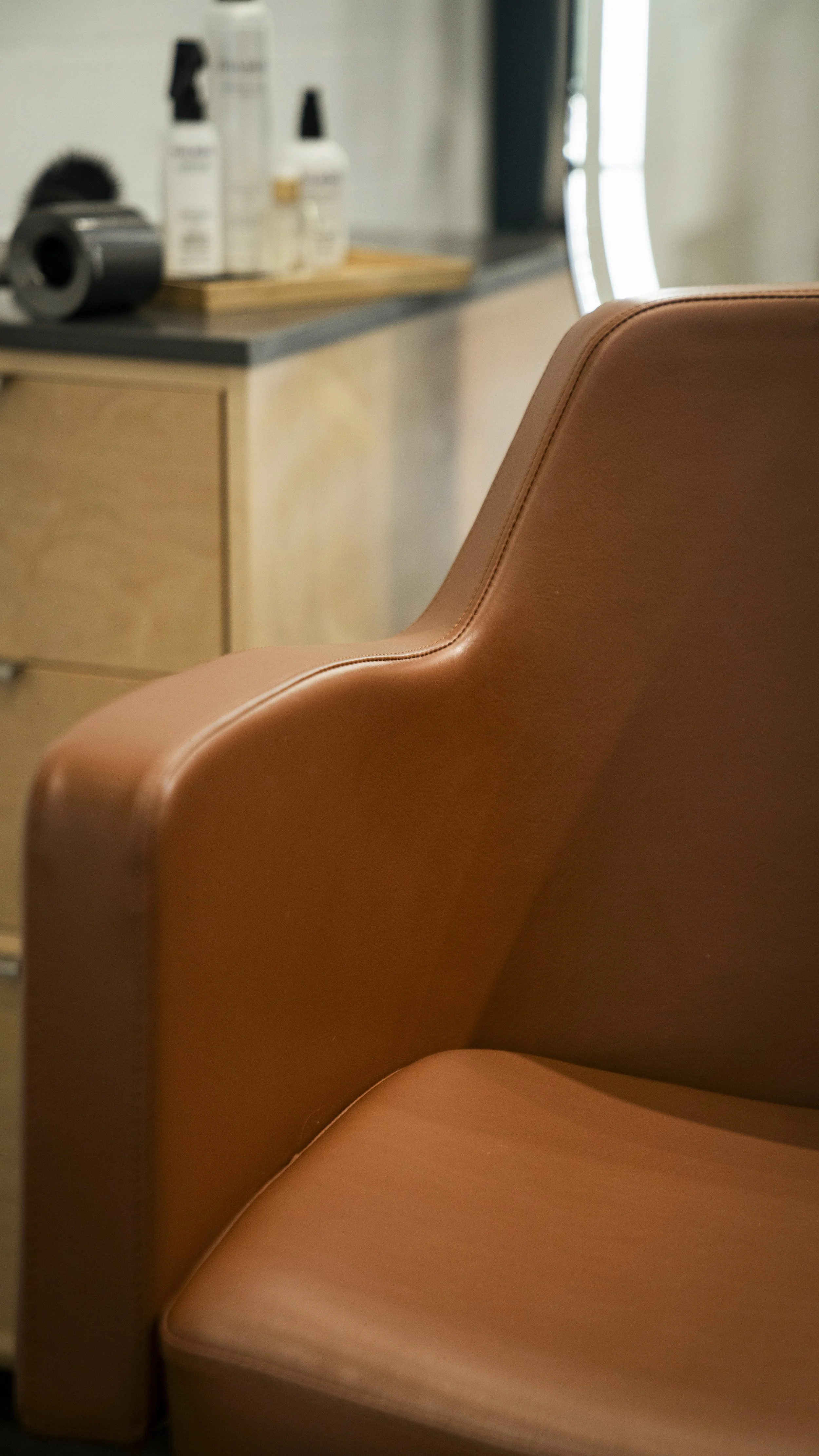 Close-up of a brown leather salon chair in a salon with a wooden cabinet and hair products in the background.