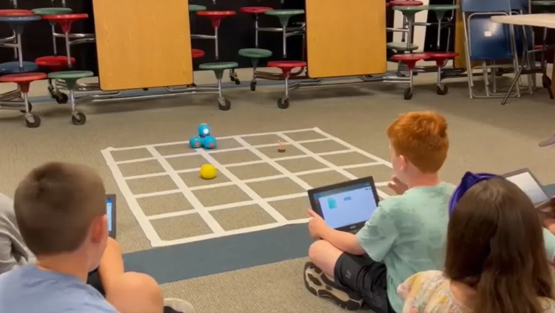 Children sitting on the floor using tablets to control a small blue robot on a taped grid layout in a classroom.