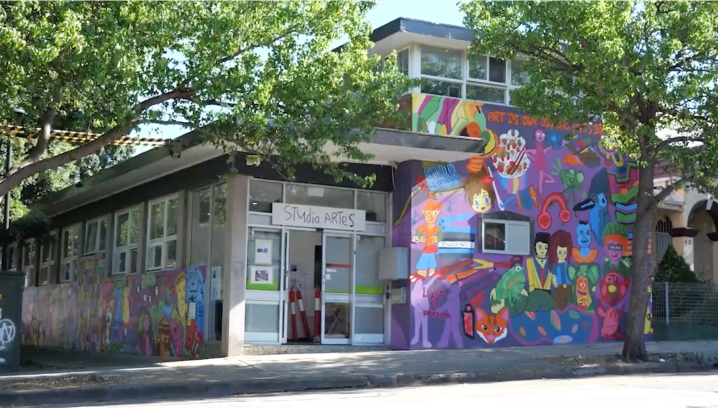 Studio surrounded by greenery, on the building itself are the words "Studio ARTES" as well as a purple mural with numerous colourful people and icons painted upon it