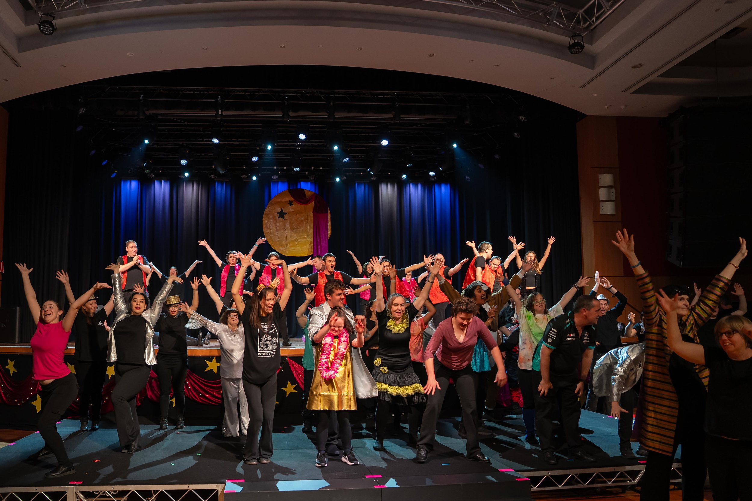 Stage filled with people in different colourful costumes performing a dance routine with their arms raised in the air, lots of smiles and stage lights shining brightly