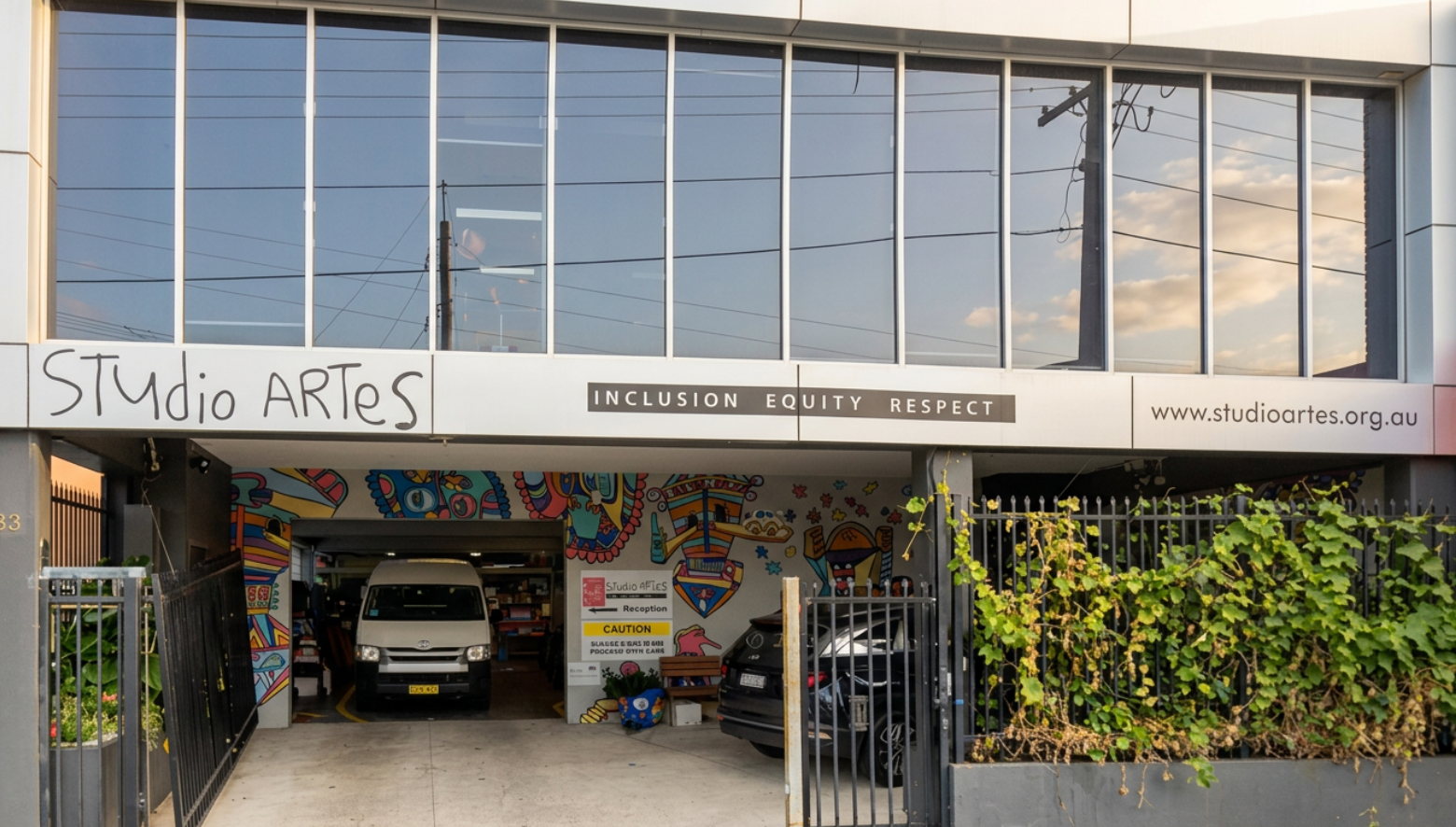 Building that has Studio ARTES in big font, next to it in smaller font is INCLUSION EQUITY RESPECT and www.studioartes.org.au. Garage space as well as green vines on the fencing and a colourful mural.