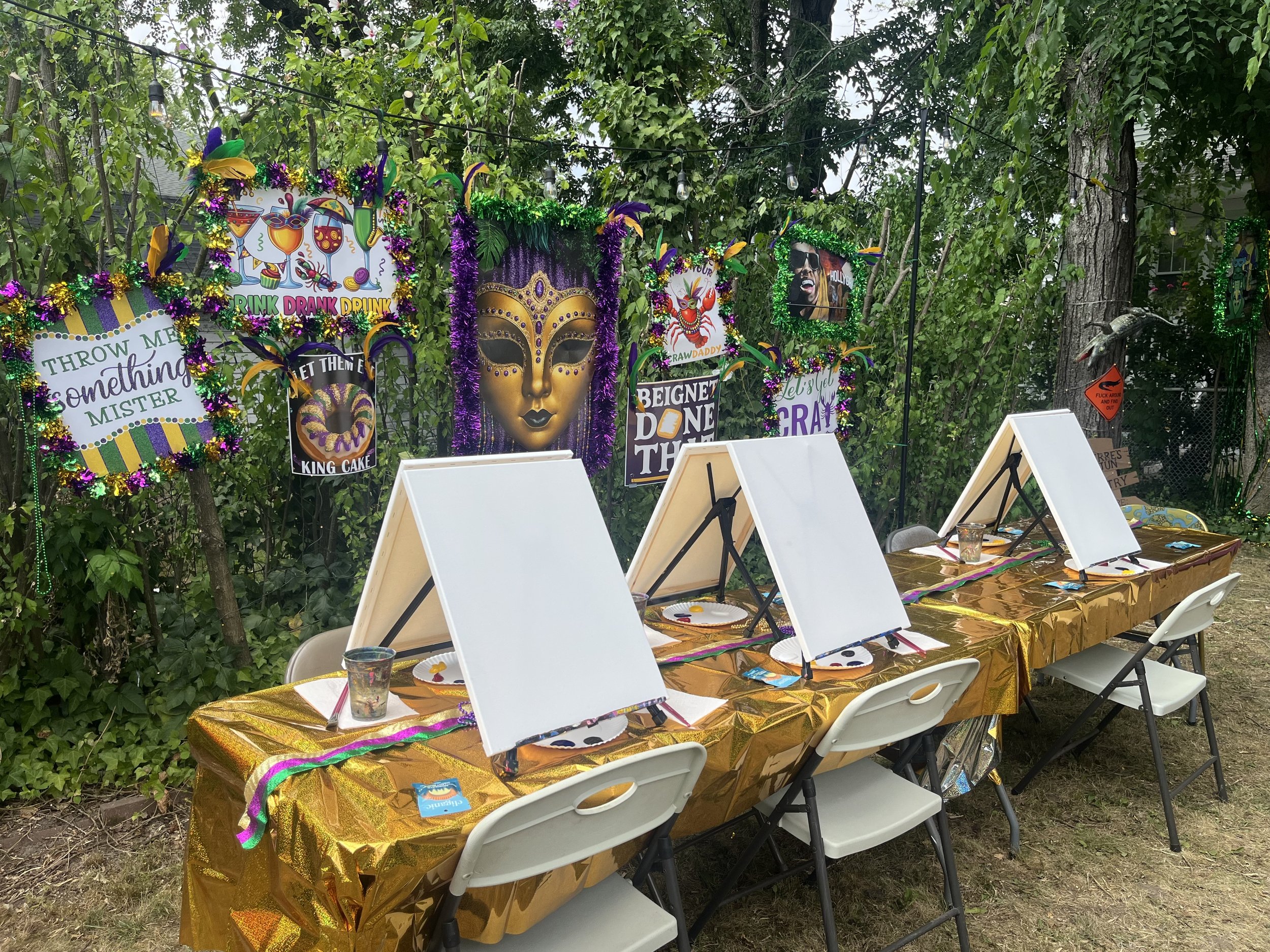 Outdoor party setup with a long table covered in a shiny gold tablecloth, decorated with colorful Mardi Gras beads, plates, and drinking cups. Behind the table are white folding chairs, and in the background, there are vibrant Mardi Gras masks and colorful decorations hanging on trees and string lights among green foliage.