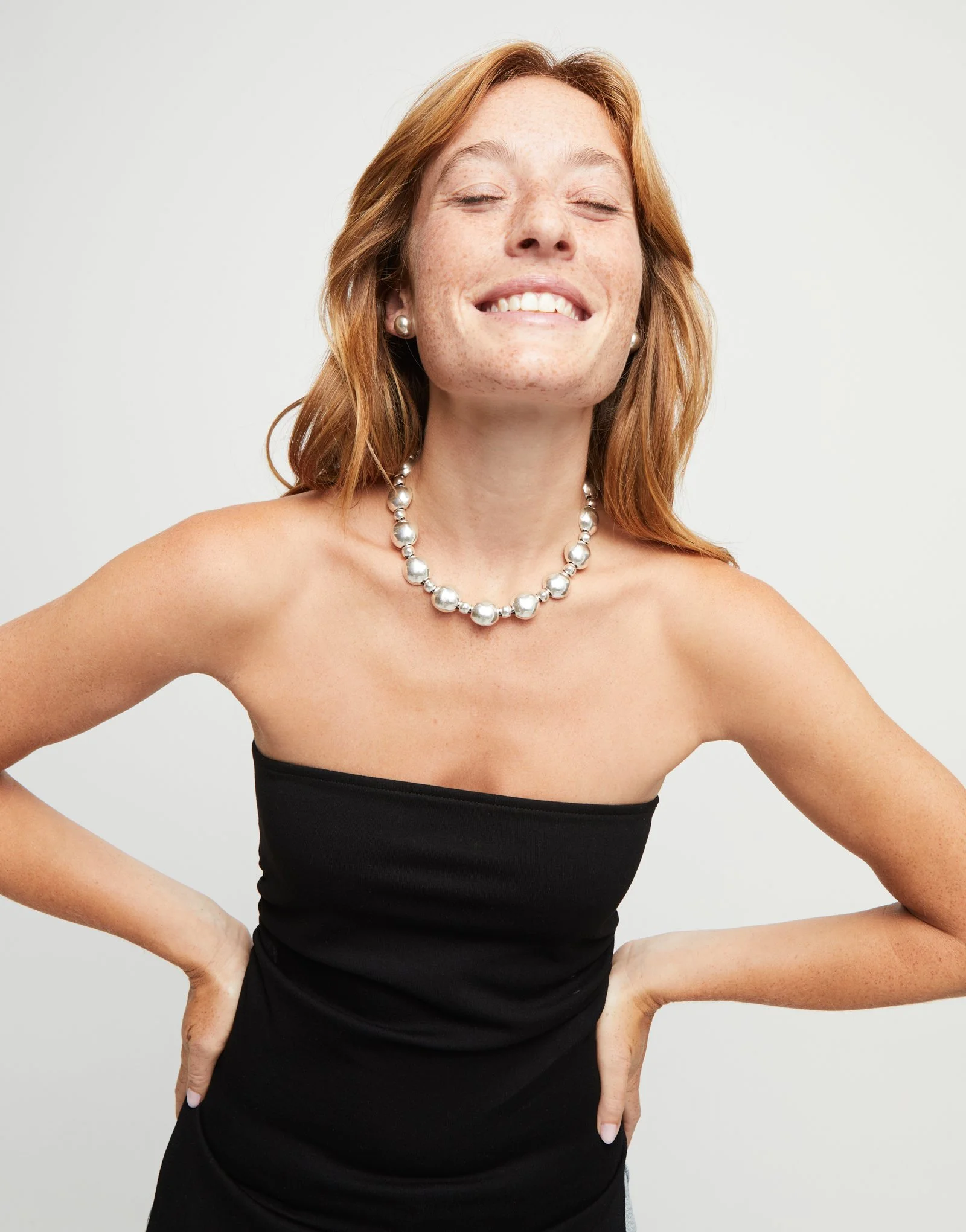 A woman with red hair and pearl earrings smiling with her eyes closed, wearing a black strapless top, and standing against a plain light gray background.