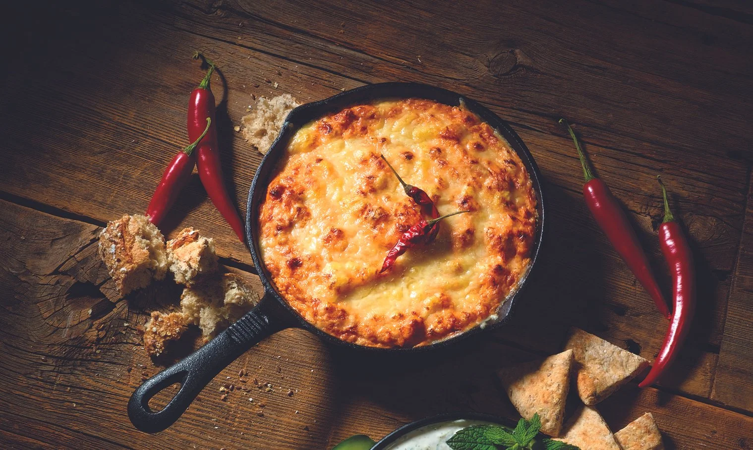 A cast iron skillet of baked cheesy dip with red chili peppers inside, surrounded by torn bread pieces, red chili peppers, and flatbread on a wooden table.
