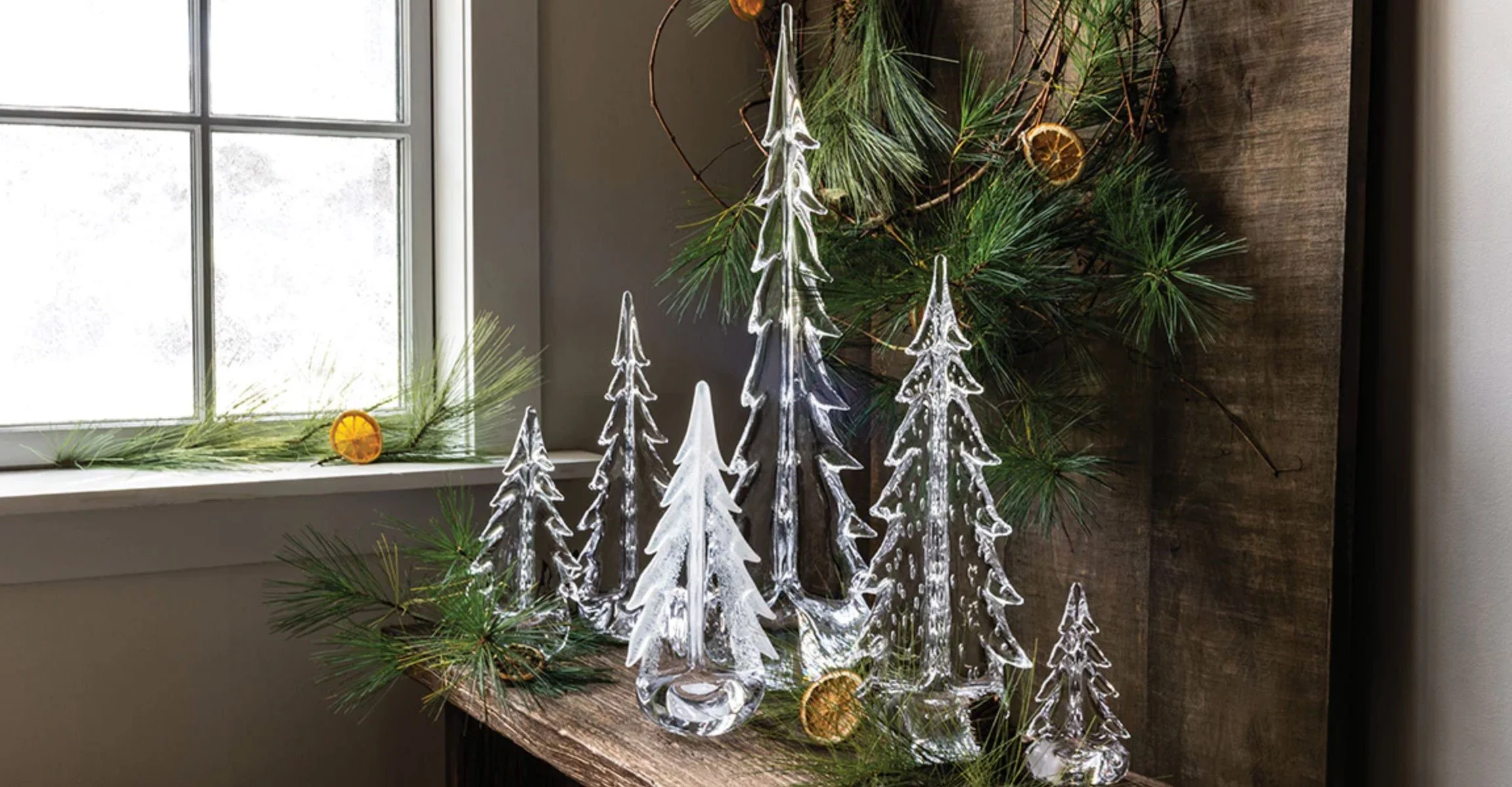 Decorative glass trees with pine branches and dried oranges on a wooden shelf near a window.