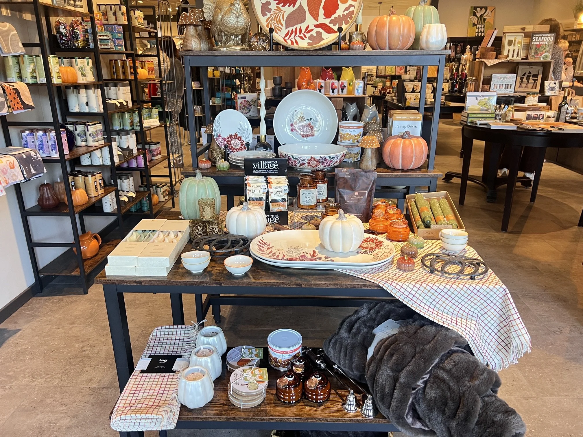 Table display with Pacific Northwest Seafood Cookbook, fish-shaped ceramic plates, wooden bowls, bowls with fish patterns, decorative dishcloths, soap dispensers, and straw baskets. Assortment of kitchenware and home goods.