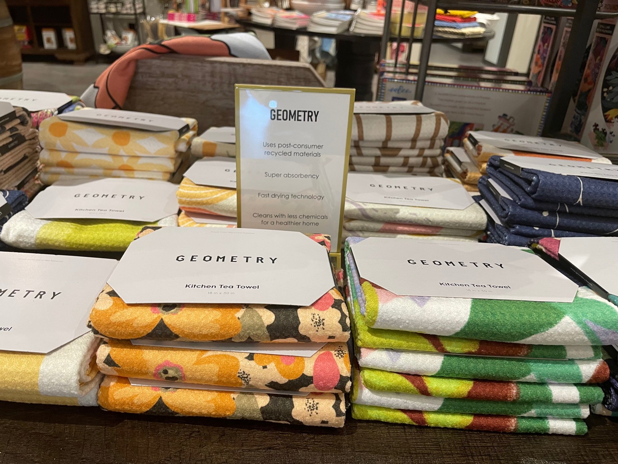Display of Geometry kitchen tea towels in various colors and patterns, stacked on a wooden table, with a promotional sign highlighting features such as absorbency and eco-friendly materials.