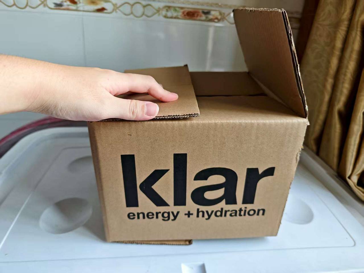 A cardboard box labeled 'klar energy + hydration' is being opened, with a person's hand lifting the flap. The box is on a white surface in a room with patterned wallpaper and curtains.