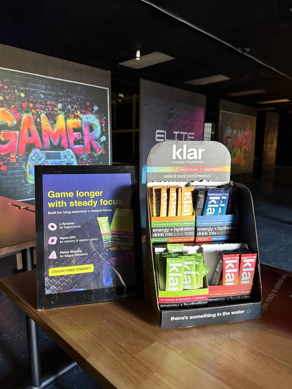 Display stand with packets of energy and hydration drink mix, including flavors like pineapple and mango, on a wooden table at a gaming event, with colorful banners and posters in the background.