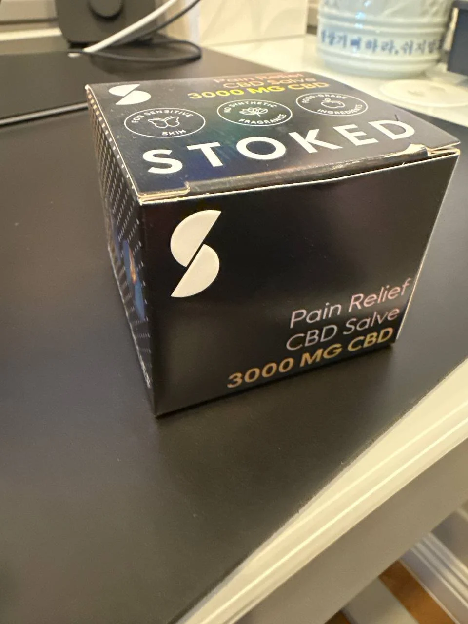 Black box labeled 'Stoked' with white logo and gold text, containing pain relief CBD salve with 3000 mg CBD, placed on a black surface.