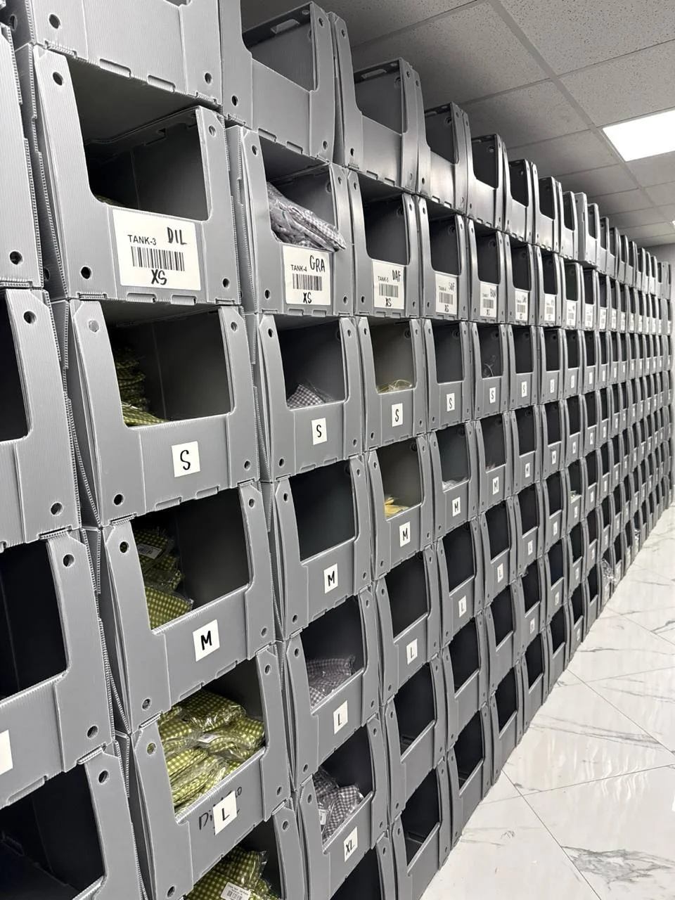 Rows of gray metal storage bins labeled with size and other labels, mounted on a wall, some bins contain small packaged items, in an organized storage room or warehouse.