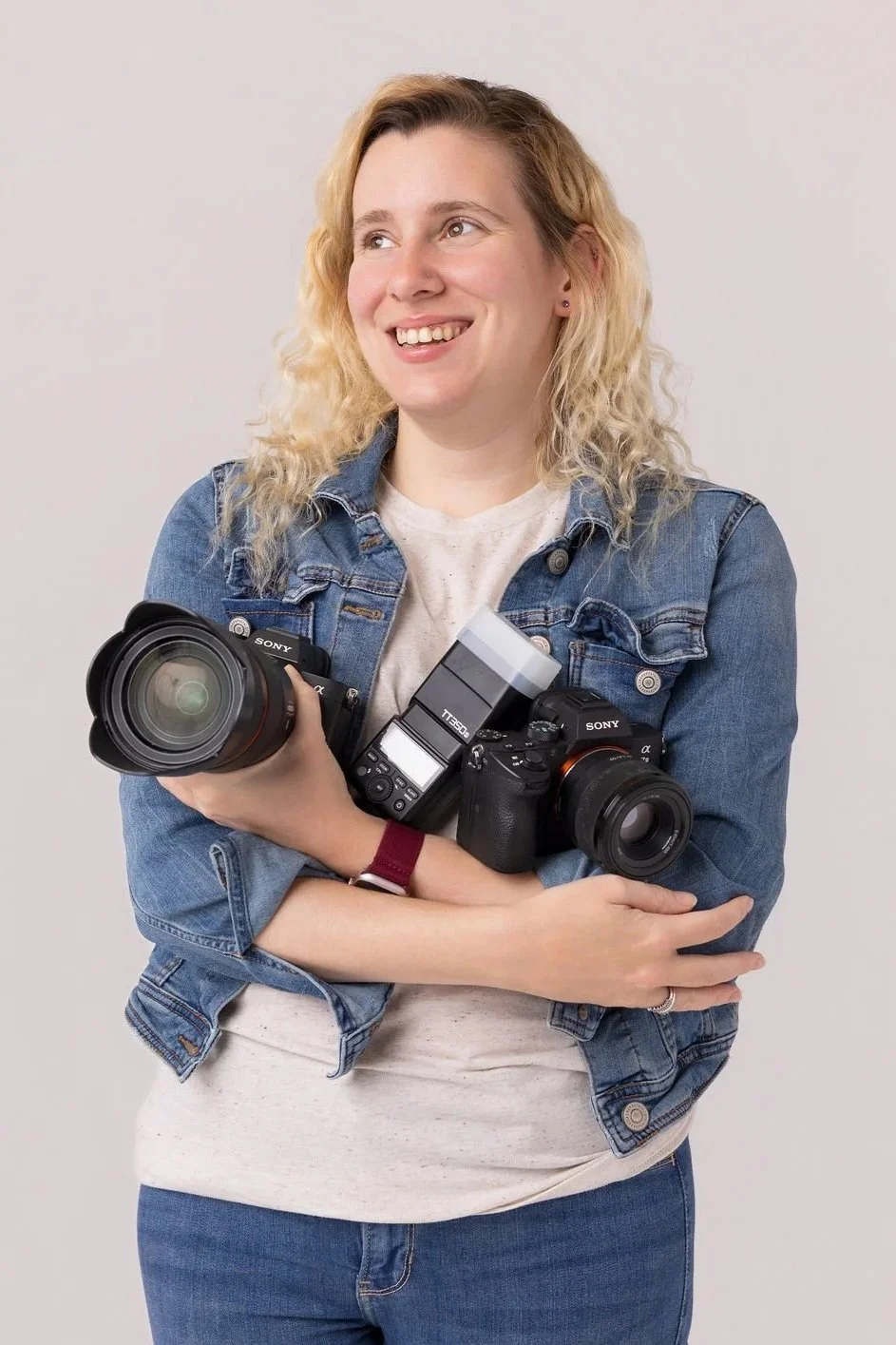 A woman with blonde curly hair wearing a denim jacket, smiling while holding two professional cameras in front of a plain light-colored background.