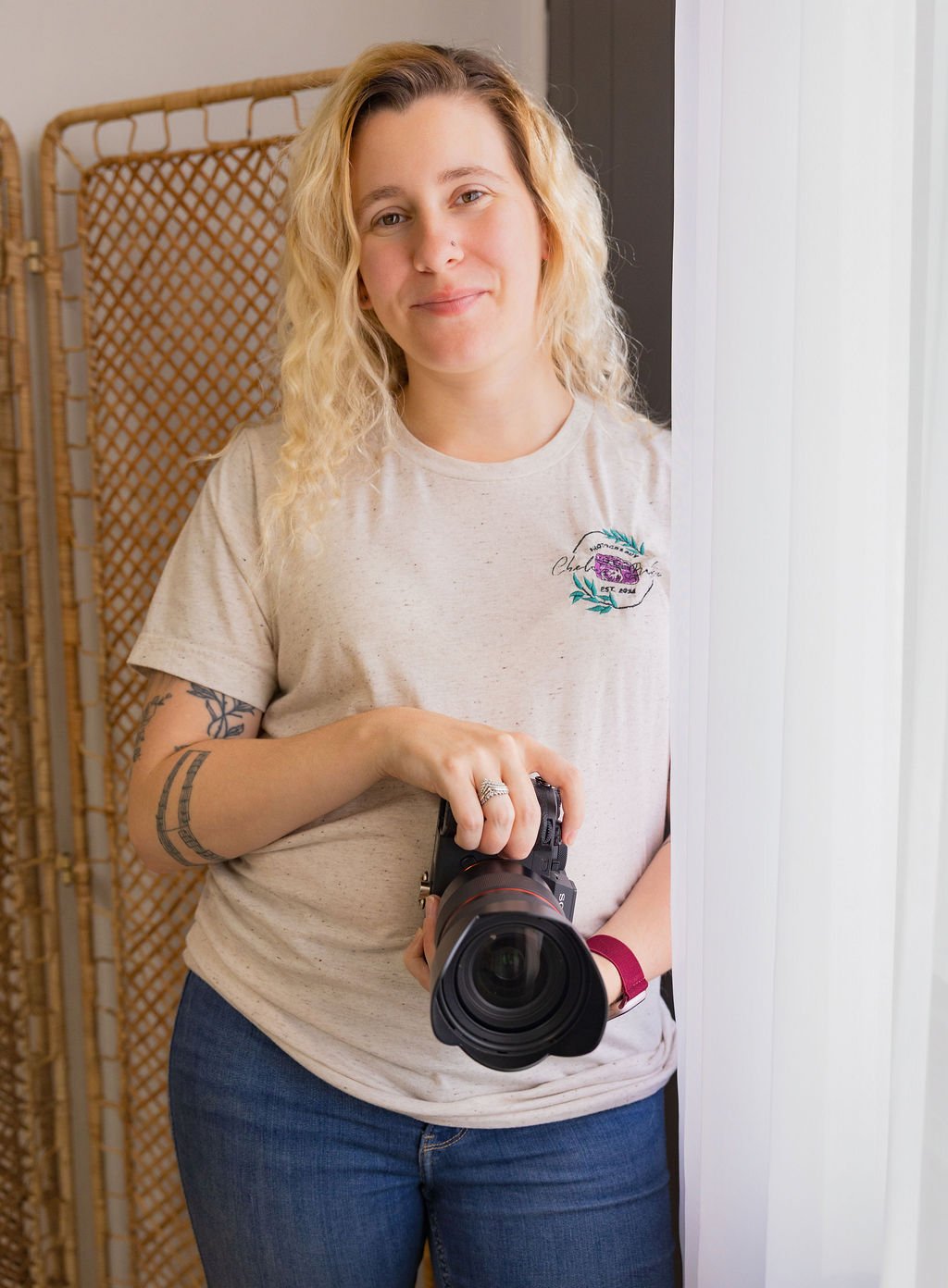 A woman with blond curly hair holding a camera, standing next to a white curtain and a woven room divider