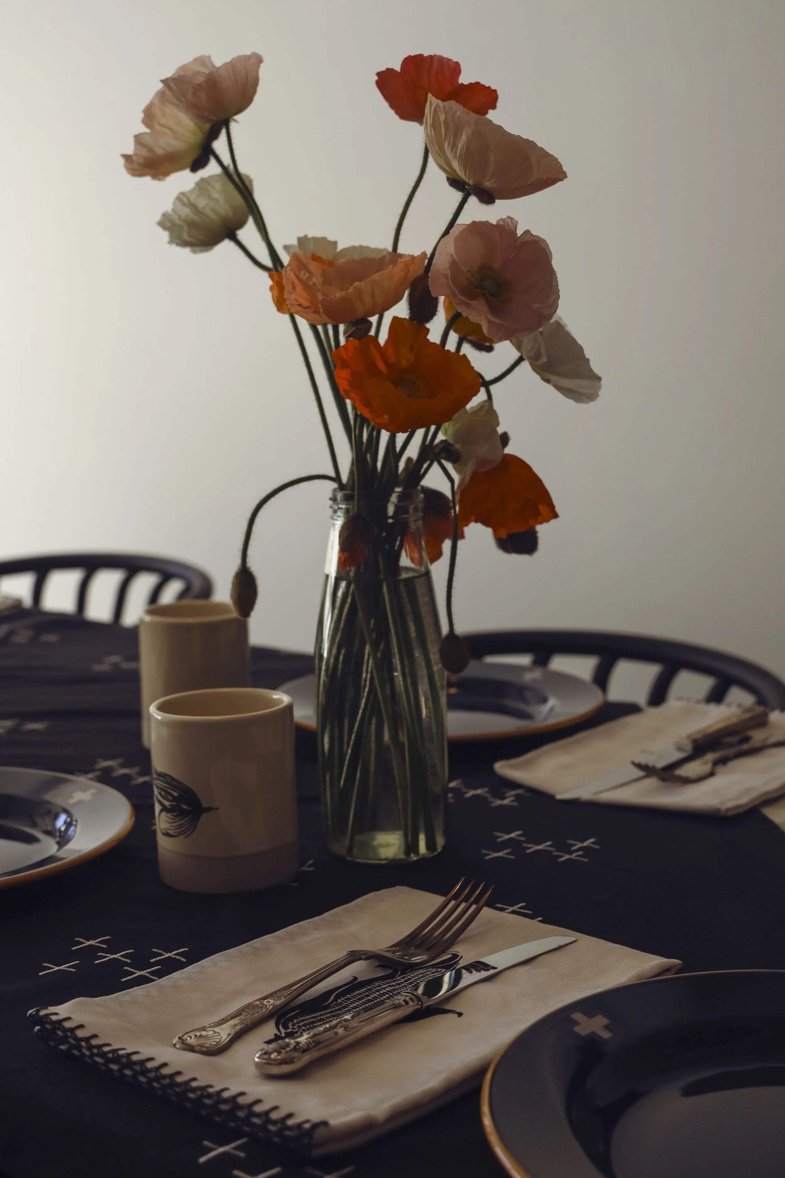 A dinner table set with black plates with gold rims, silver cutlery on white napkins, and a tall glass vase with pressed flower bouquet of pink, orange, and cream-colored flowers. There are two white cups, a black chair, and a tiered serving tray in the background.